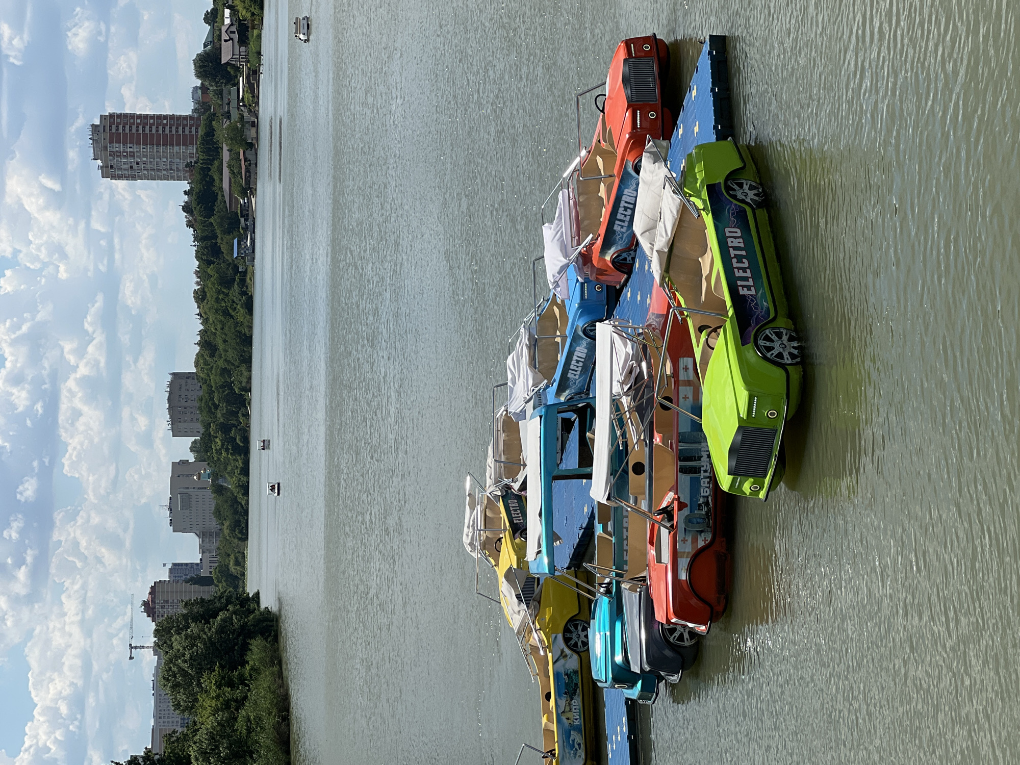 This image shows a row of colorful, car-shaped pedal boats docked on a calm body of water, likely a river or lake, on a sunny day. The boats are various shades of green, red, orange, and blue, and each has "ELECTRO" written on its side in a stylized font with lightning bolt accents. Some boats have white covers over their seating areas. The boats are lined up in a parallel fashion, suggesting they are available for rent.
In the background, a lush green treeline runs along the shore, behind which several multi-story buildings and a tall apartment building are visible. A construction crane is also present, indicating ongoing development in the area. The sky is partly cloudy with fluffy white clouds against a blue backdrop. Two small boats with people in them are visible further out on the water. The scene appears to be in Krasnodar, Russia, suggested by the urban architecture and the presence of the city's characteristic apartment buildings. The overall mood is relaxed and recreational, indicative of a pleasant day for outdoor activities.