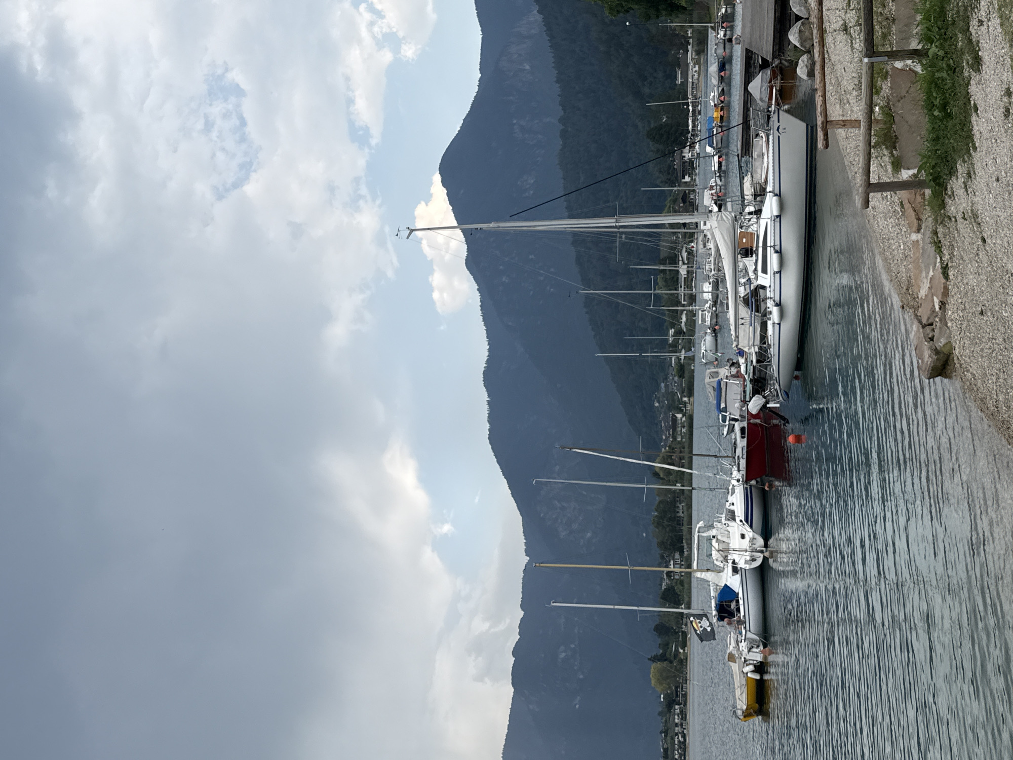 The image shows a body of water with several sailboats docked along a shore. The water is calm, reflecting the overcast sky and the dark green mountains in the background. A small cluster of buildings and trees are visible along the shoreline, nestled at the base of the mountains. The foreground features a gravelly beach with a rustic wooden fence. The scene appears to be a quiet harbor, possibly in a mountainous region.
