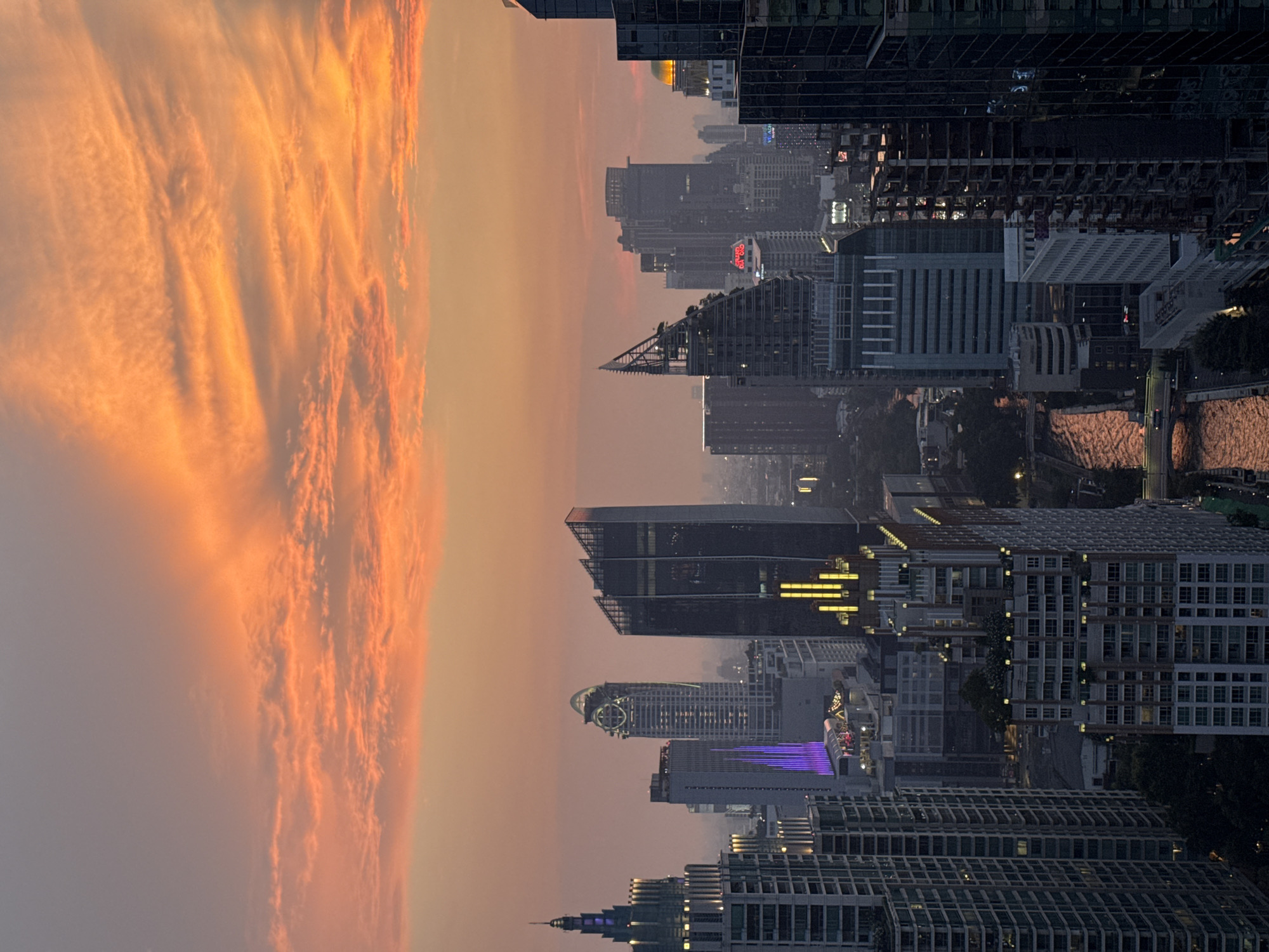 The image shows a cityscape at dusk, likely in Bangkok, Thailand, given the architectural style and the presence of skyscrapers. The left side of the image is dominated by a sky filled with orange and pink clouds, indicative of sunset or sunrise. On the right, densely packed buildings and skyscrapers with lit windows suggest a bustling urban environment. Streetlights and building illumination contribute to a dynamic and somewhat hazy atmosphere. A river or waterway reflecting ambient light is visible between some of the structures. The overall impression is one of a vibrant city merging with a colorful sky.