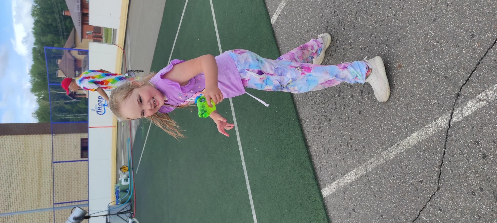 The image shows a young girl with blonde hair wearing a purple t-shirt and colorful floral patterned pants. She is holding a green toy gun in her right hand and appears to be smiling. She is standing on an asphalt surface with white painted lines, likely a sports court. In the background, there is a green sports field with a white border, a brick building, and trees. A person wearing a red cap and a tie-dye shirt is visible in the distance. The scene suggests an outdoor event or activity in Noviy, Russia.