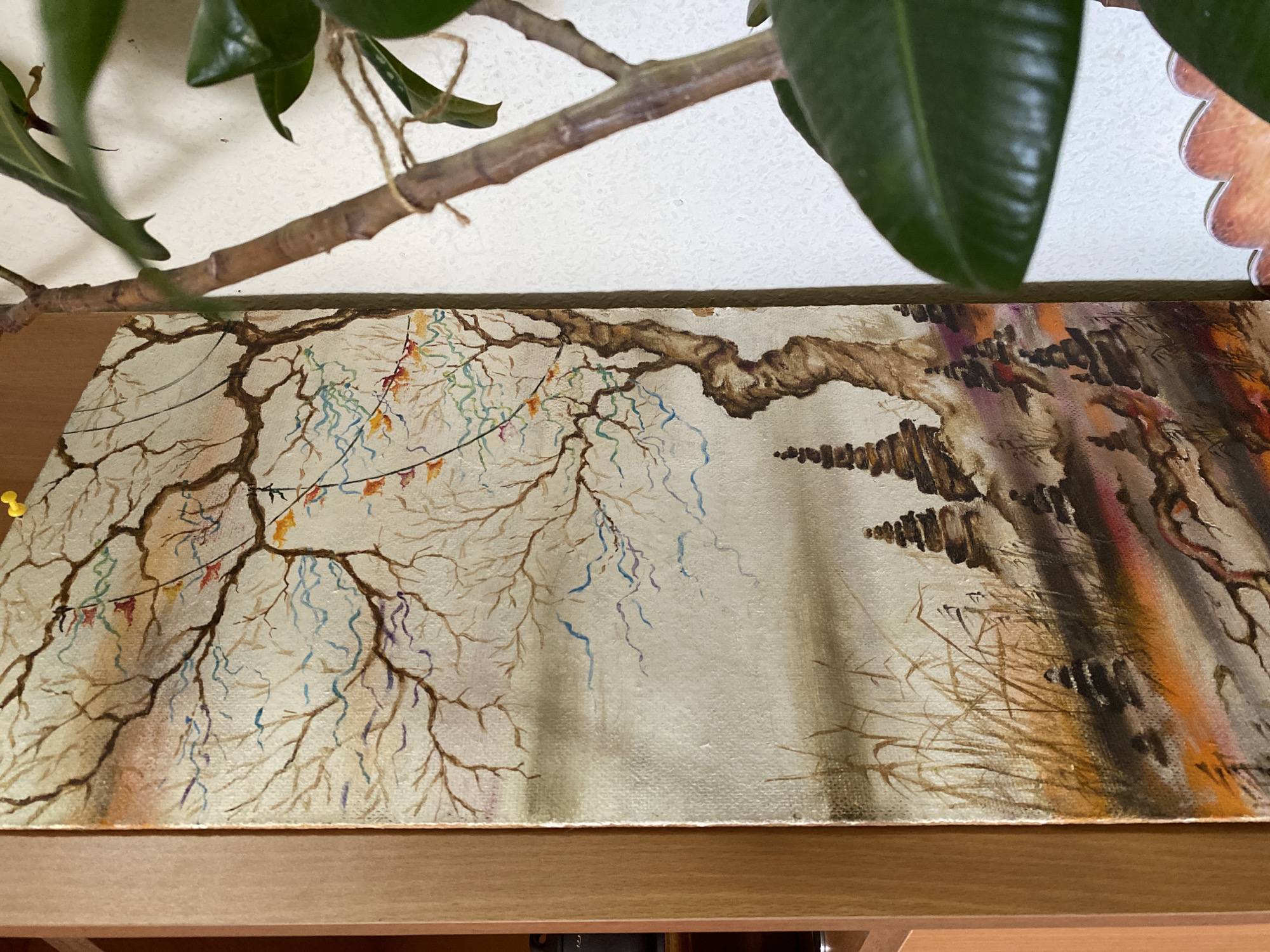 A close-up shot of an unframed painting, featuring a bare tree with decorations, likely streamers and small flags, reminiscent of a festive occasion. The right side of the painting includes a less distinct, possibly wooded area with vertical markings. The painting is resting on a wooden surface, likely a shelf or ledge. Partially obscuring the top of the image is a houseplant with thick, waxy leaves and a slender branch. The background consists of a white wall. There are no people visible in the shot. The location in Chaikovsky, Russia cannot be verified from the image.