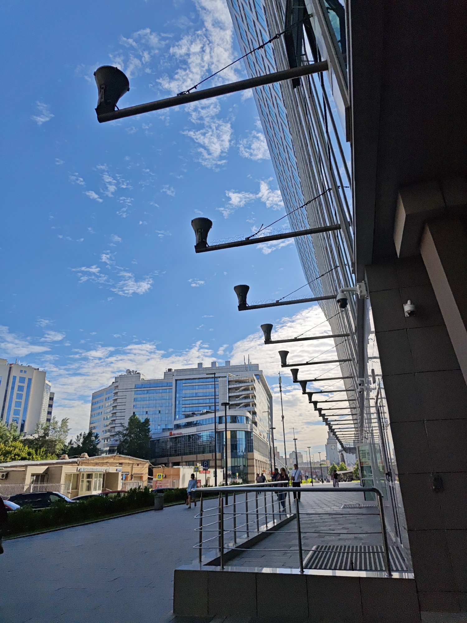 The image captures a street scene in Moscow, Russia, with a focus on modern architecture and urban infrastructure. The foreground features a sidewalk with a metal railing, leading towards a contemporary building with a glass facade. Several identical, dark-colored speakers are mounted on horizontal metal poles extending from the building's exterior, with a few security cameras also visible. In the background, other tall, modern buildings with glass windows are prominent under a bright blue sky dotted with white clouds. A few trees and smaller structures are also present, along with a parked car. Several people are seen walking on the sidewalk, either alone or in small groups, suggesting a typical urban daytime environment. The overall impression is one of a well-developed, modern cityscape.