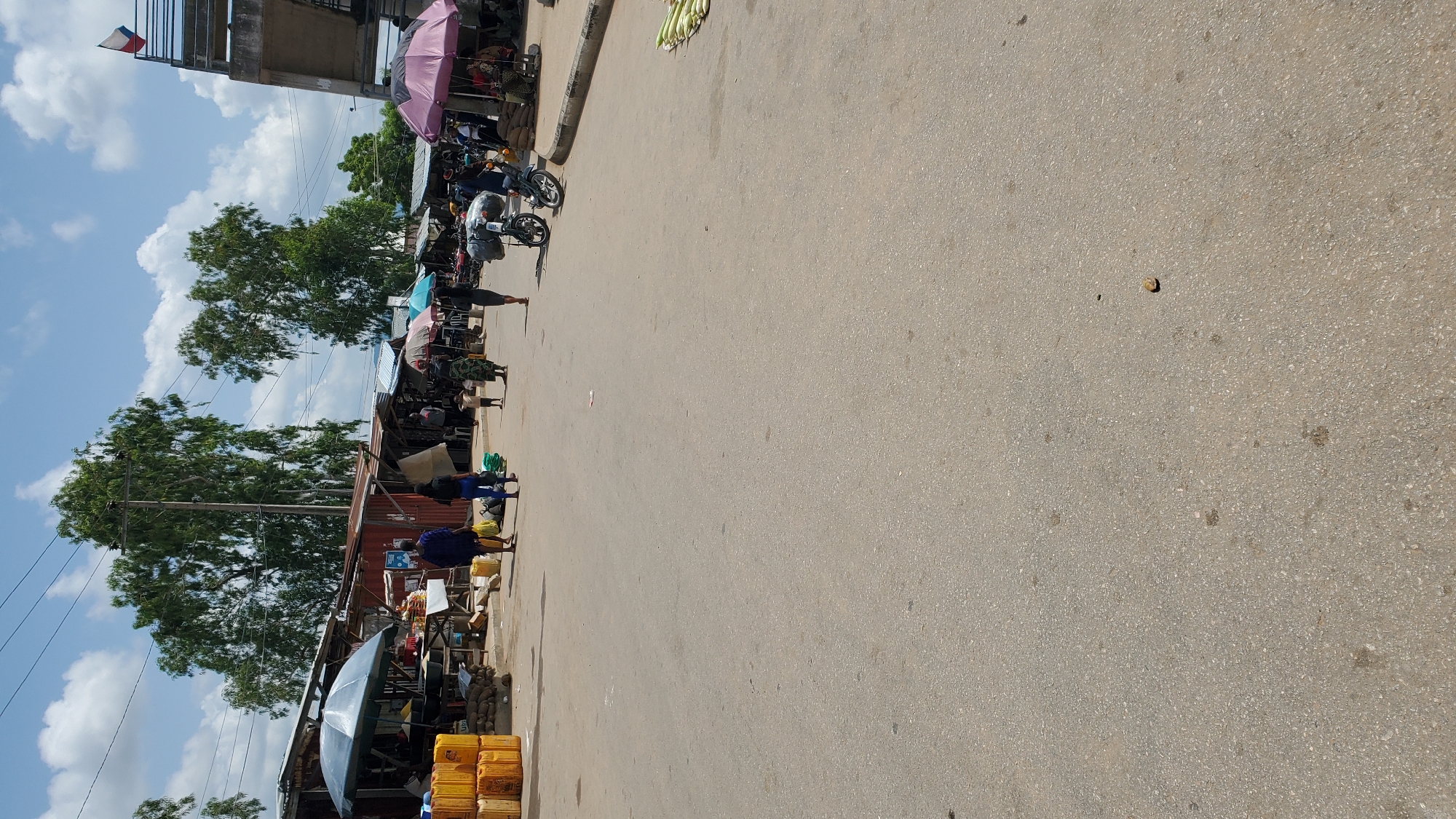 The image shows an outdoor market scene in Jalingo, Nigeria. The foreground is an asphalt road, with a few scattered debris. In the mid-ground, there are market stalls set up along the side of the road, with various goods on display. Several people are walking through the market. To the left, there are stacks of yellow plastic containers. Several umbrellas are open, providing shade for some stalls. In the background, there are trees and buildings, and a pedestrian overpass can be seen on the right side of the image, with a flag at its top. The sky is blue with scattered clouds.