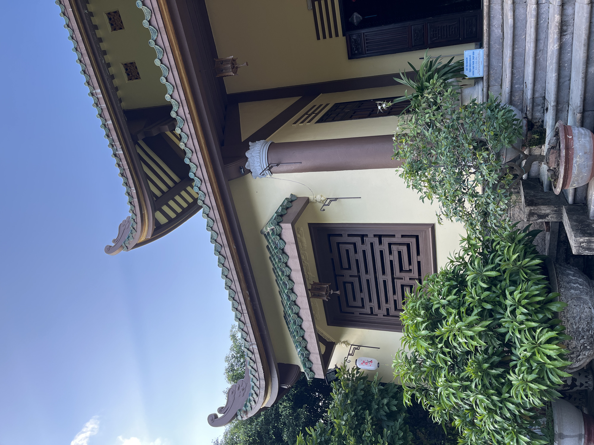 The image captures a portion of a building in Đà Nẵng, Vietnam. The building features a traditional Asian architectural style with a curved roof adorned with green tiles. A brown post supports the roof structure, and there is a window with an intricate geometric design. Potted plants are placed near the building's entrance, adding greenery to the scene. A small sign with Vietnamese script is visible near the steps, likely providing information or instructions. The sky is clear and blue, suggesting a bright, sunny day.