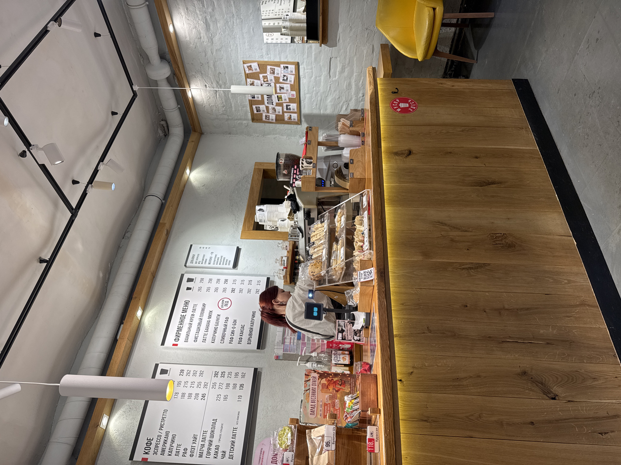 The image depicts the interior of a café, likely in Saint Petersburg, Russia based on the Cyrillic text on the menus and signs.
A woman with red hair is standing behind a wooden counter. She appears to be working, perhaps as a barista or server. Behind her is a display case filled with pastries and other baked goods.
The café has a modern, rustic design with white brick walls and exposed pipes on the ceiling. There are menus displayed on the wall listing various coffee drinks, including "espresso," "Americano," "cappuccino," and "latte," along with other beverages and pastries.
A yellow chair is visible near the counter. A small bulletin board with photos or notes is affixed to the wall. A sign reading "Дом" (Home) is visible. Prices for the items are listed in rubles.