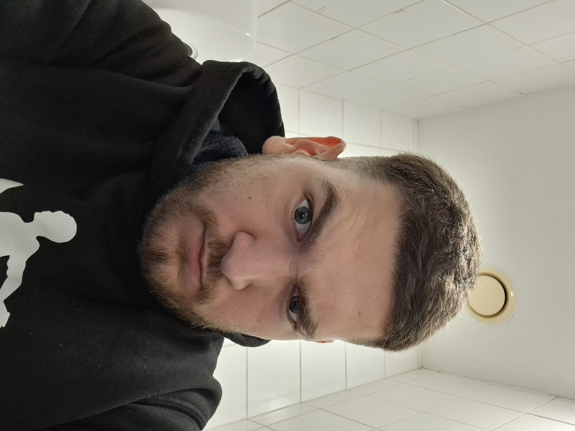 A close-up, low-angle selfie shows a man in a black hoodie with a white graphic print. He has short brown hair, a beard, and dark eyes. His eyebrows are furrowed, giving him an angry expression. He is looking directly at the camera. The background is a white tiled wall with a ventilation fan in the ceiling. The scene appears to be a bathroom, possibly in Nicosia, Cyprus, given the tile style which is common in older buildings in the region.