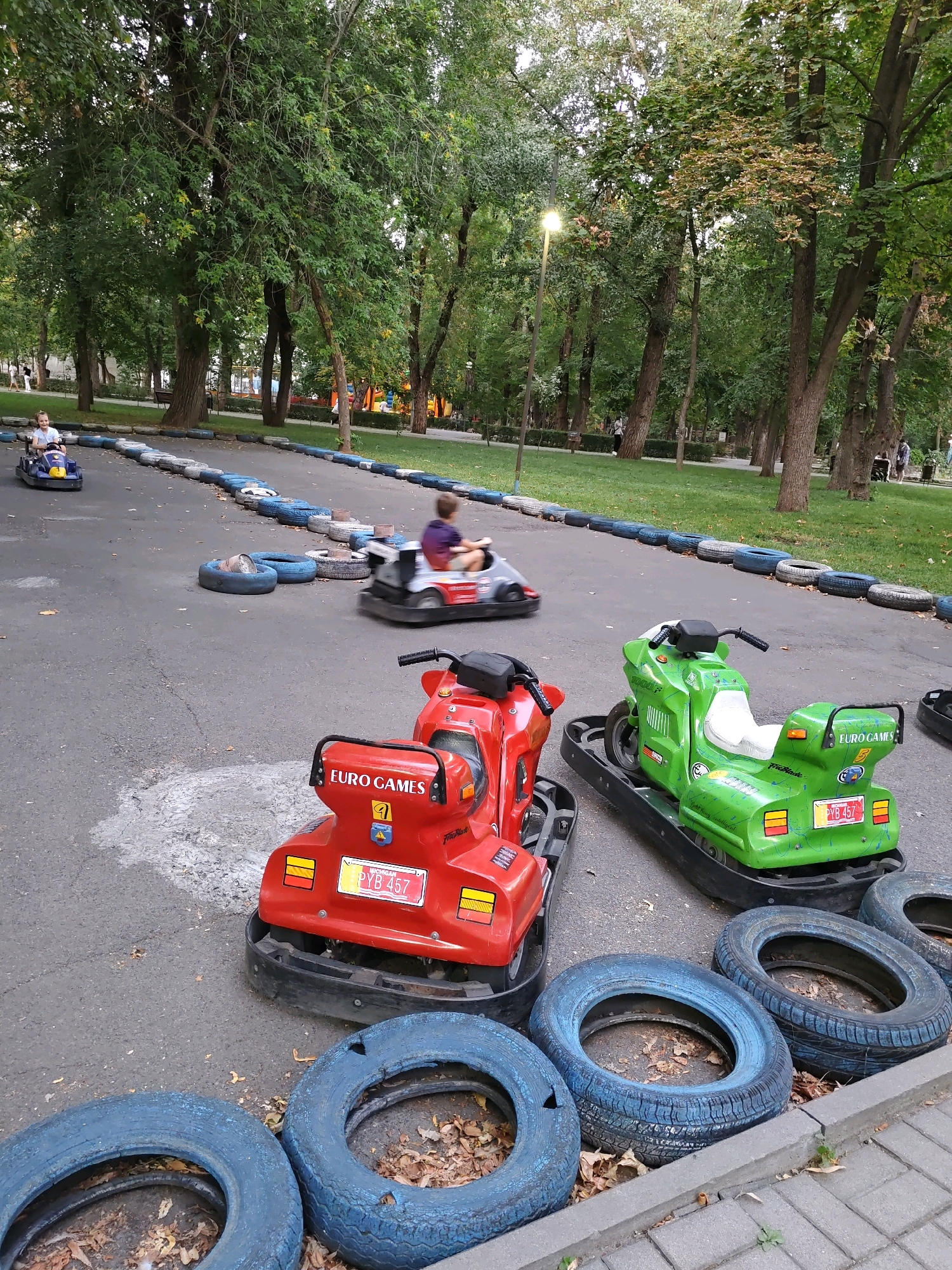The image depicts a go-kart track in a park setting, likely in Rostov-on-Don, Russia. Several go-karts, including a red, a green, and a gray one, are visible on the asphalt track, which is bordered by tires. One go-kart has the text "EURO GAMES" and a license plate that reads "PYB 457". A child is driving the gray go-kart, and another child is visible in a blue go-kart further down the track. In the background, trees and a lamppost suggest an outdoor park environment.