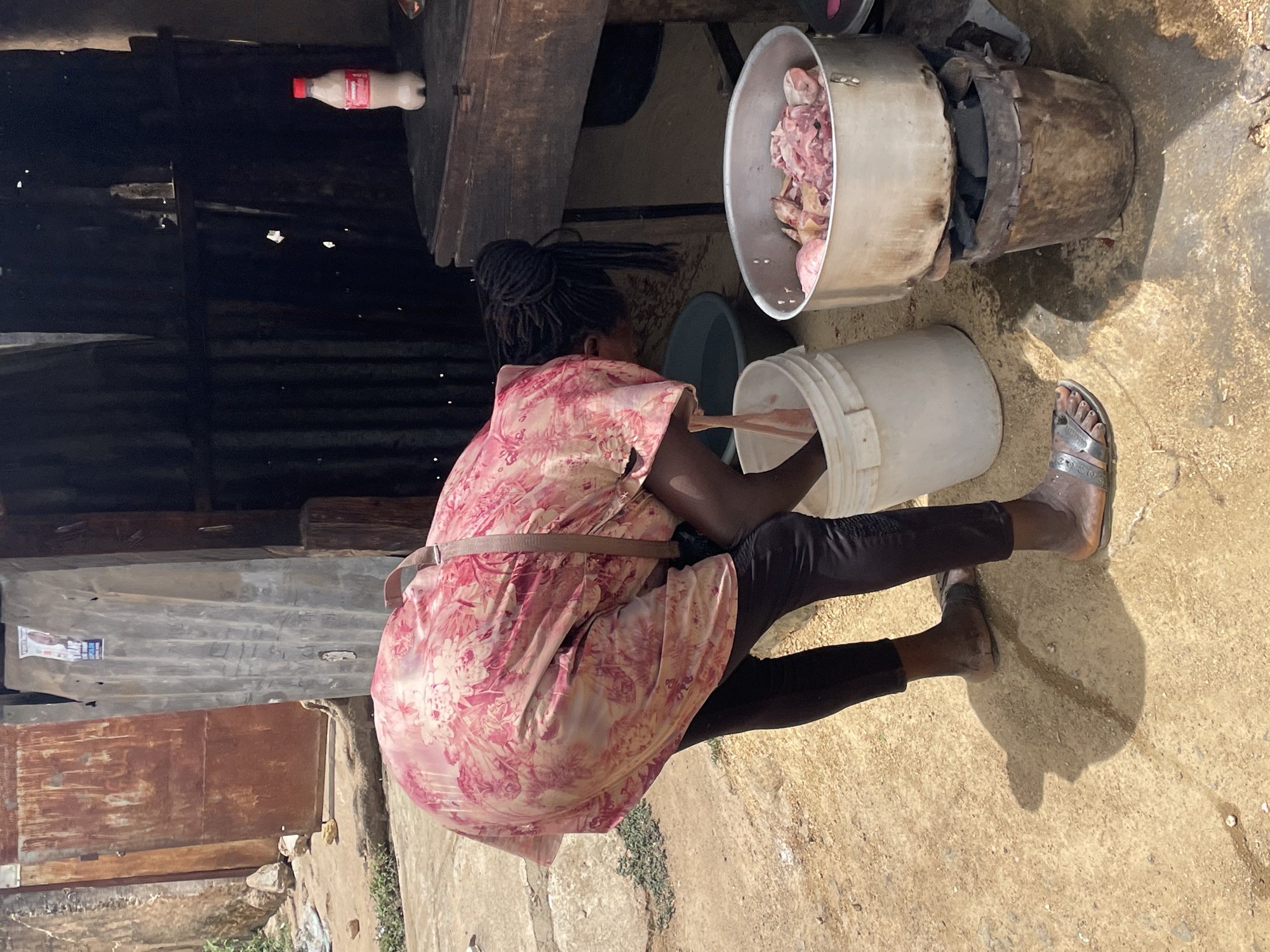 A woman is squatting in an outdoor area. She is wearing a floral patterned pink and white dress, dark leggings, and silver sandals. Her hair is styled in braids. She appears to be transferring food from a large metal pot into a white plastic bucket. The metal pot is sitting on a makeshift stove constructed from a smaller metal container containing charcoal. Behind her is a rudimentary structure with corrugated metal walls and a wooden table. A plastic bottle is resting on the table. The ground is concrete. The location is likely Jos, Nigeria.