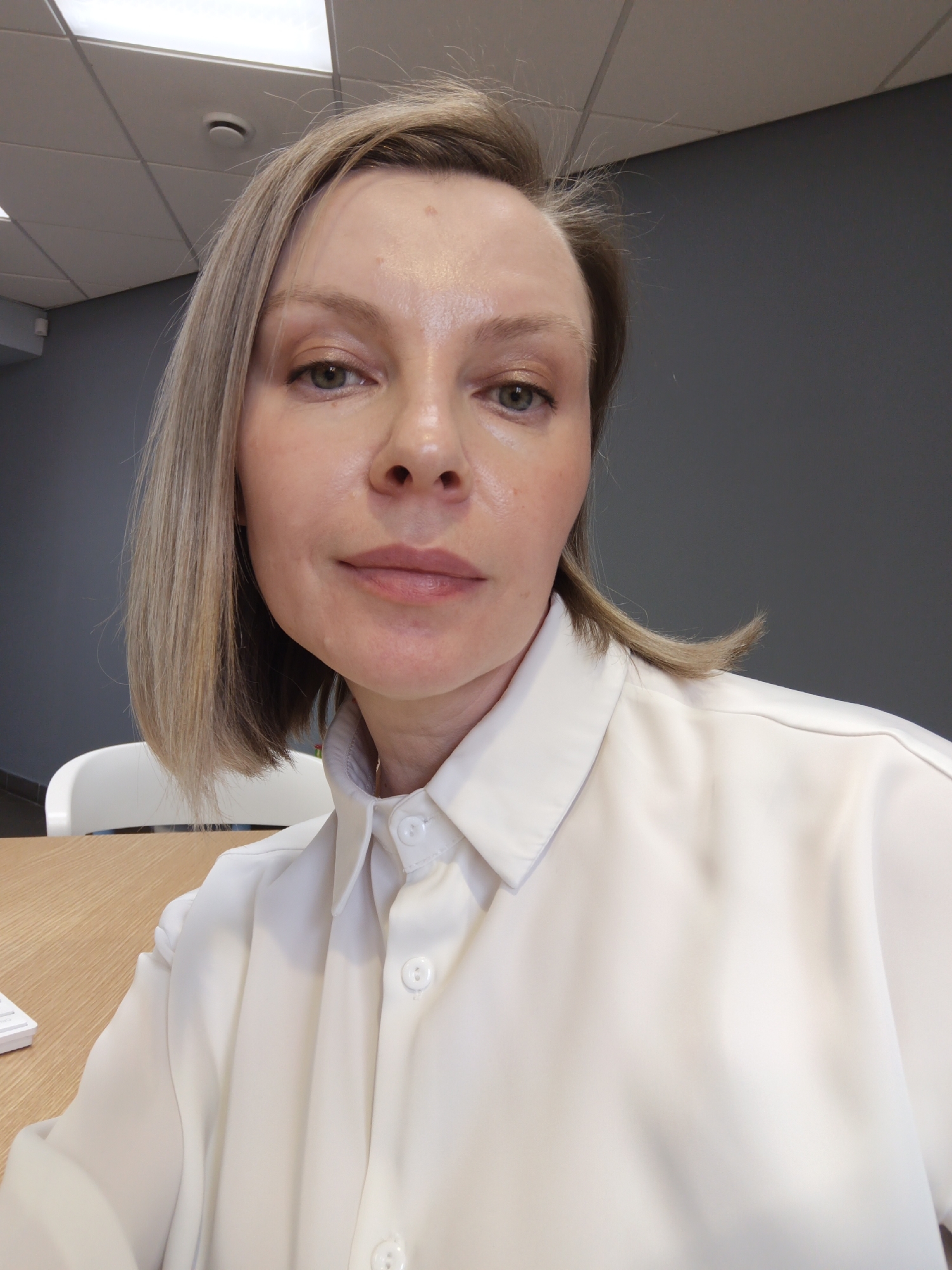 A head-and-shoulders portrait features a woman indoors, likely in an office setting in Chaikovsky, Russia. The woman, appearing to be in her 30s or 40s, has fair skin, light brown or blonde shoulder-length hair, and light-colored eyes, possibly greenish-grey. She is looking directly at the camera with a neutral, calm expression. She wears a light-colored, possibly white or off-white, collared button-down shirt.
The background indicates a professional environment. A dark grey wall is visible behind her, topped by a white ceiling with acoustic tiles and integrated fluorescent lighting. A white smoke detector and a white security camera are noticeable on the ceiling and a distant wall, respectively. Partially visible to her left is a white office chair, and a light-colored wooden desk or table is in the foreground, with a small white object on it. The lighting is bright and even, characteristic of indoor daytime office illumination. No visible text is present in the image.