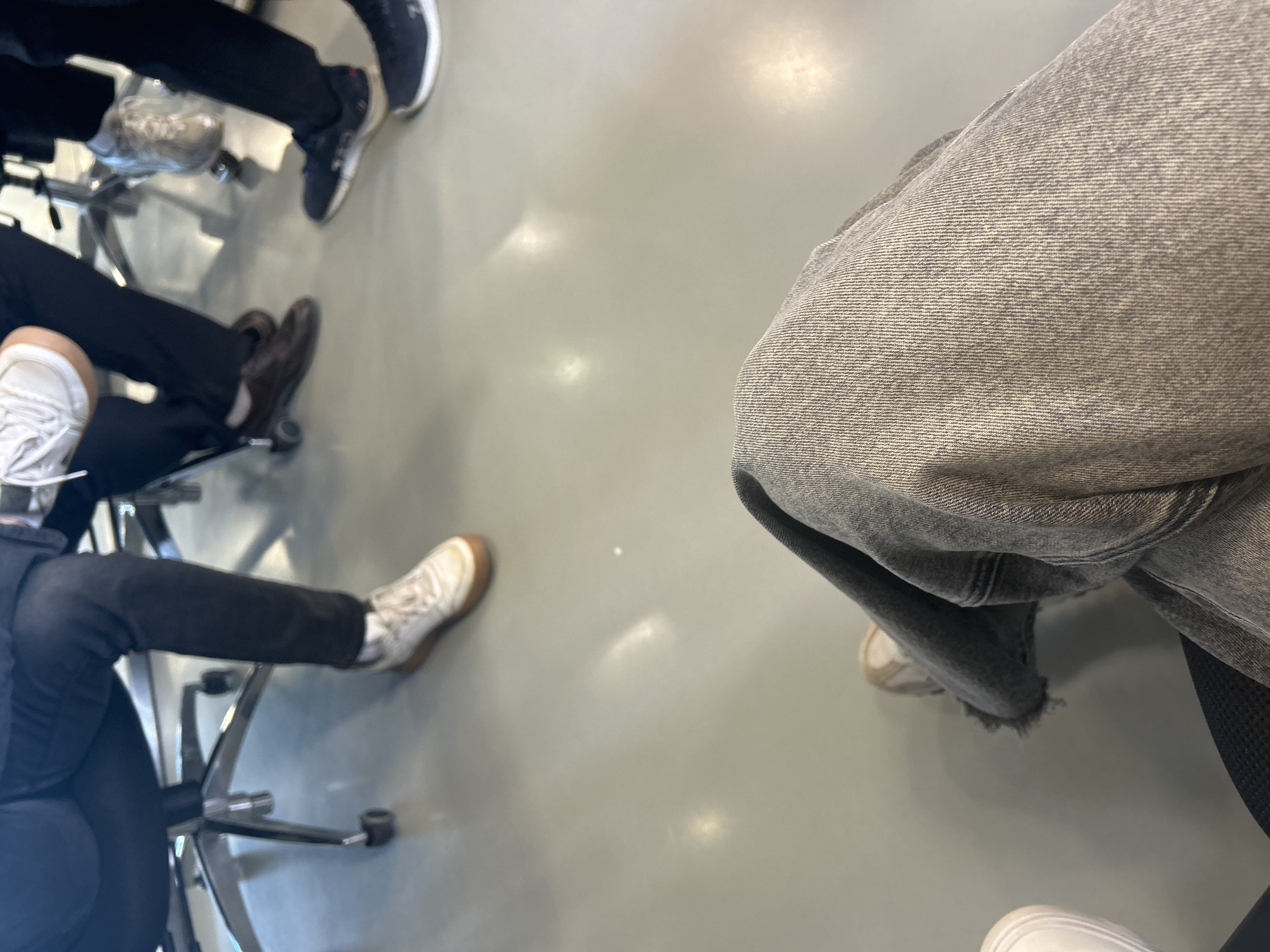 In this medium-shot, eye-level, indoor photograph, the perspective is from a person seated on a rolling office chair, looking down at their own legs and the legs of others seated around them. The foreground is dominated by the seated person's grey, textured denim jeans, with one leg bent at the knee and the other partially visible. The person is wearing a light-colored sneaker with a brown sole.
In the mid-ground, the legs and feet of other individuals are visible. One person wears dark pants and white sneakers with brown soles. Another wears dark pants and brown leather shoes. A third person is in dark pants and dark sneakers. A fourth person's legs are mostly obscured, but they appear to be wearing dark pants and dark sneakers. The individuals are seated on office chairs with metal bases and wheels, on a smooth, light grey floor. The lighting in the scene is bright, creating reflections on the floor.
The overall impression is a group of people in a casual, indoor setting, possibly an office or meeting room. There is no visual information to suggest the specific location within the city of Perm, Russia, or any other city.