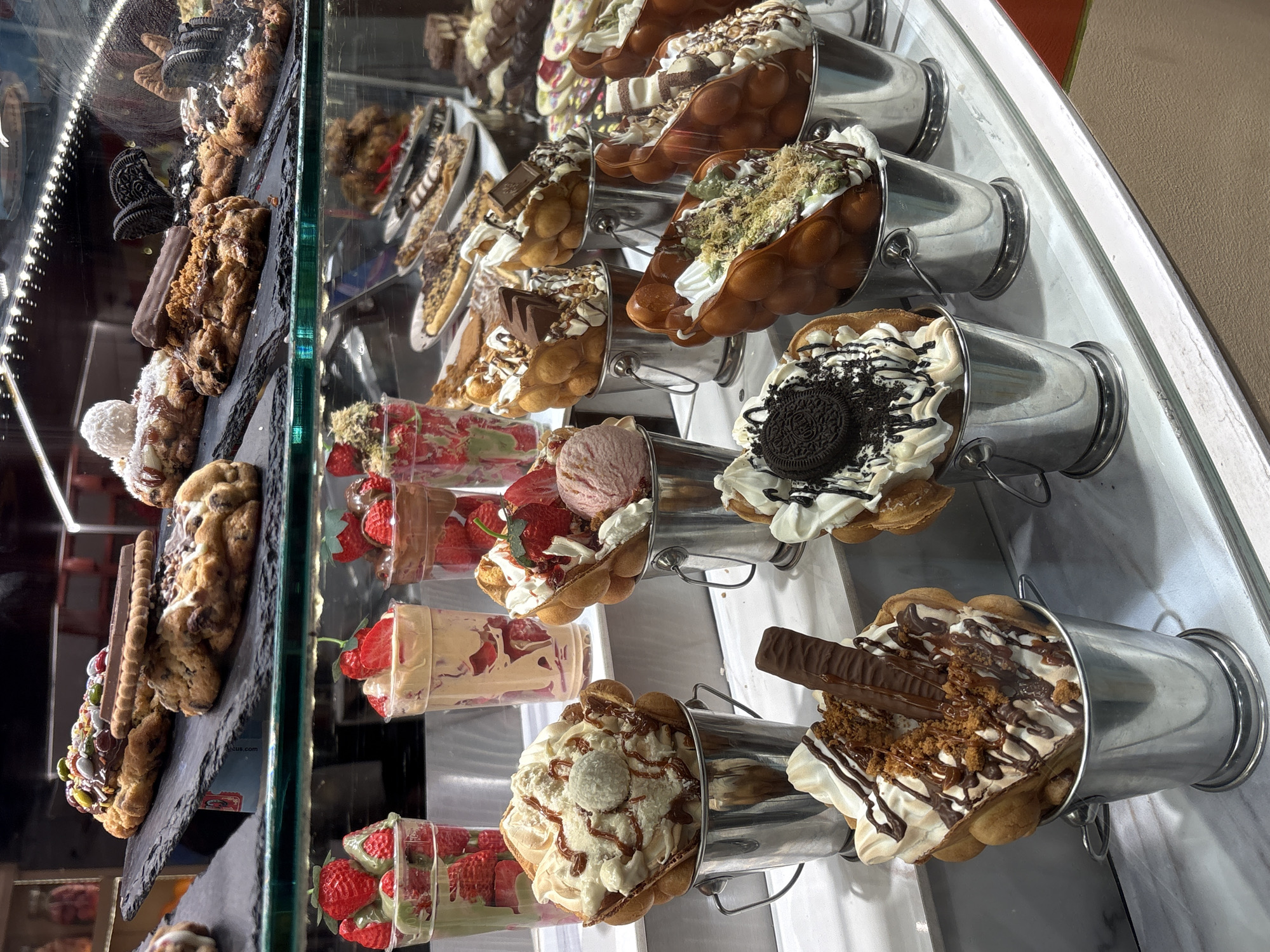 This image depicts a dessert display case in what appears to be a dessert shop or cafe. The items for sale include cookie stacks and bubble waffle desserts in metal buckets, along with cups of fresh strawberries. The presence of multiple ice cream and dessert choices suggests a focus on sweet treats. Given the prevalence of shops of this type in Barcelona, Spain, it could be reasonably assumed that the location is in Barcelona, Spain. However, definitive confirmation of the location is not possible from the image.