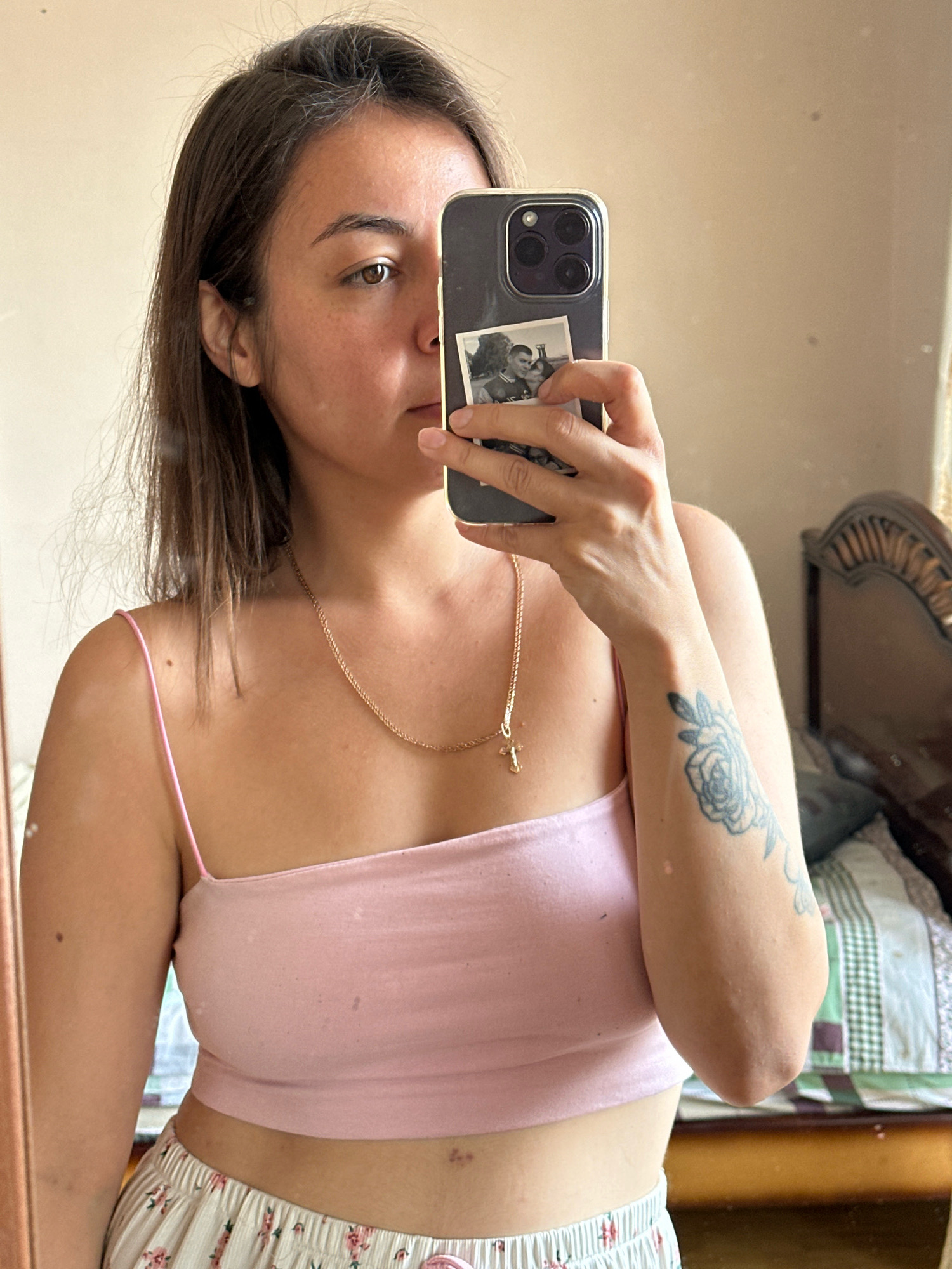 A young woman with shoulder-length brown hair takes a mirror selfie in Gyumri, Armenia. She is wearing a light pink spaghetti strap crop top and patterned pajama pants. A gold chain necklace with a cross pendant rests on her collarbone. A tattoo of a rose is visible on her left forearm. She is holding a smartphone in her right hand, which has a small black and white photo adhered to the back. The background shows a mirror, a bed with a patterned comforter, and a wooden headboard, consistent with an indoor residential setting.