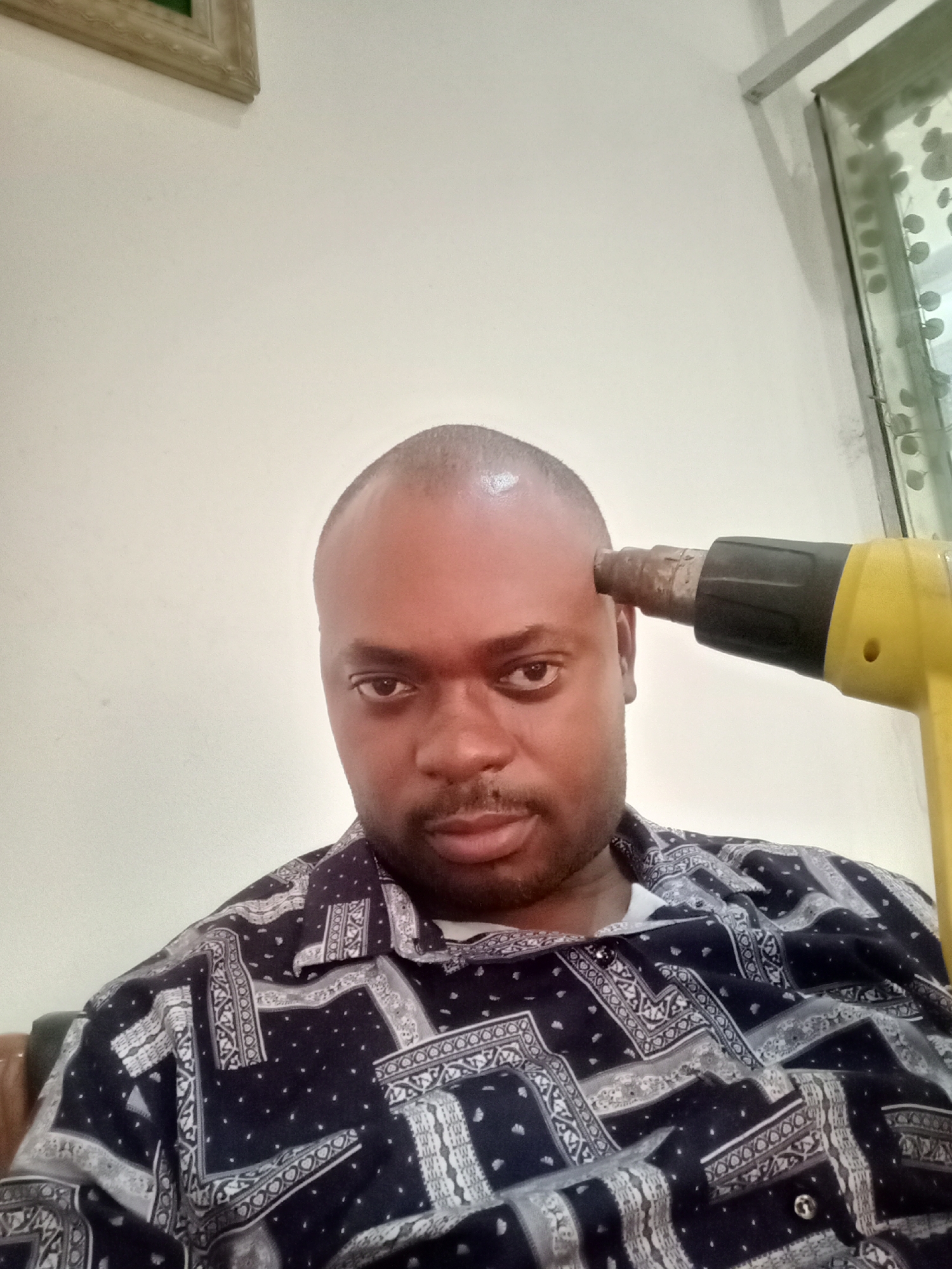 A medium shot depicts a man holding a yellow heat gun pointed at his head. The man is dark-skinned with a bald head. He wears a patterned black and white shirt. The background is a white wall, a framed picture is partially visible on the upper left, and a patterned glass window is to the upper right. The location is Gwarinpa, Nigeria.