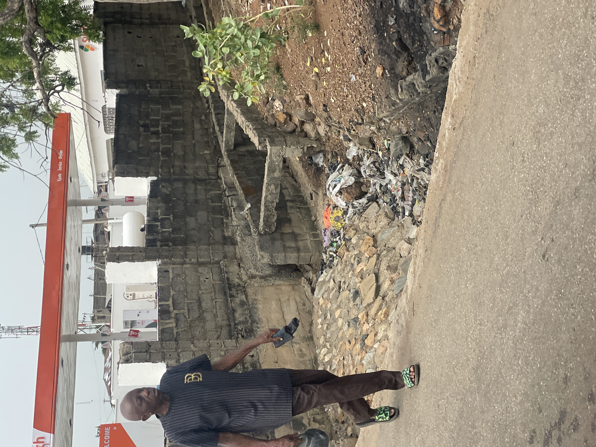 A man stands on a concrete path in Lokoja, Nigeria. He wears brown trousers, black and grey patterned shirt, and patterned sandals. He is holding a small object in his right hand and a hat in his left hand. To his right is a dilapidated brick structure with a pile of trash in the adjacent ditch. To his left, a petrol station with orange columns is visible.