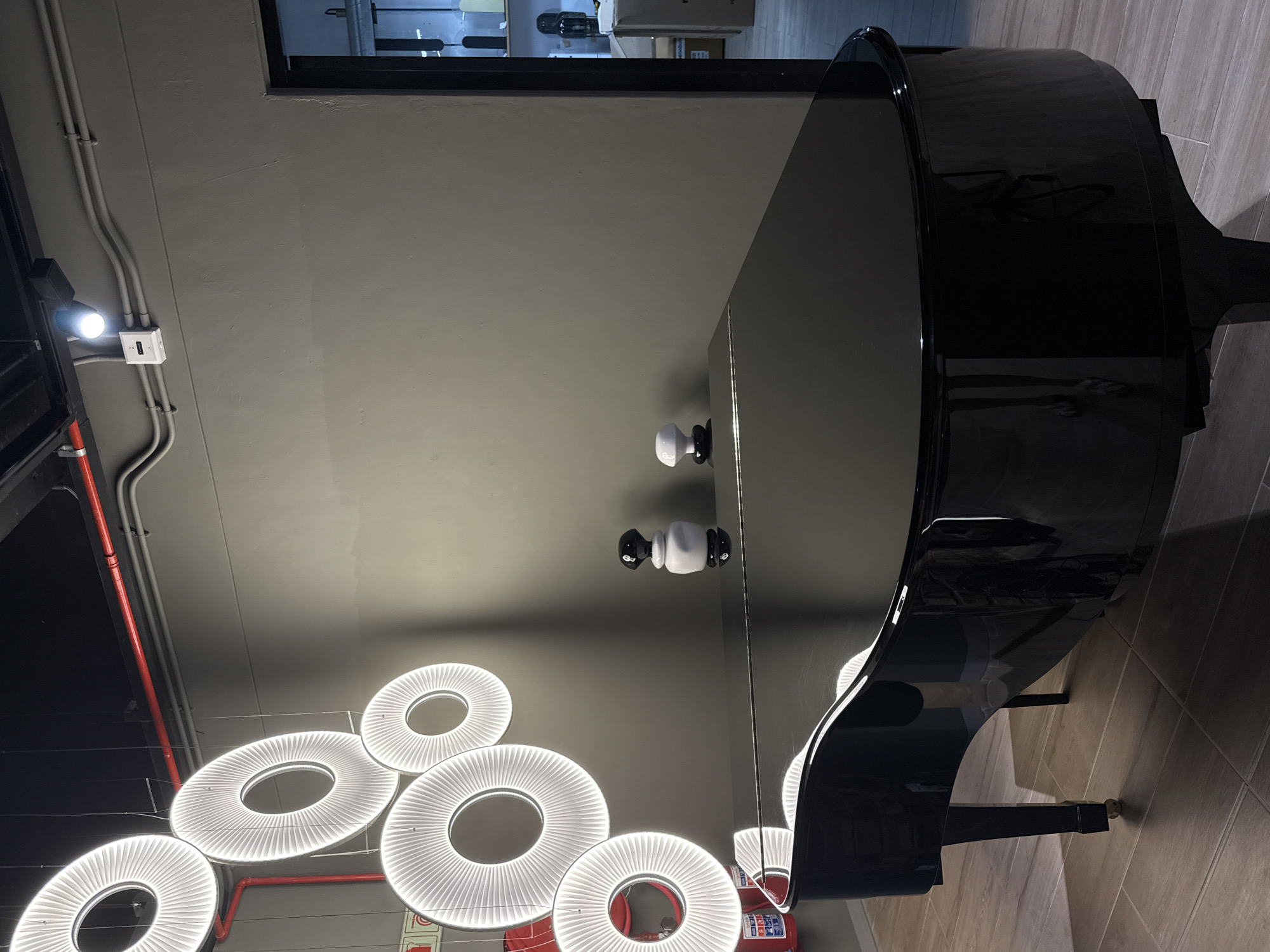 The image depicts the interior of a space in Cape Town, South Africa, featuring a polished black grand piano. Circular LED light fixtures hang from the ceiling. Electrical conduits and fire suppression equipment are visible, indicating a commercial or public setting. There are no people visible in the shot.