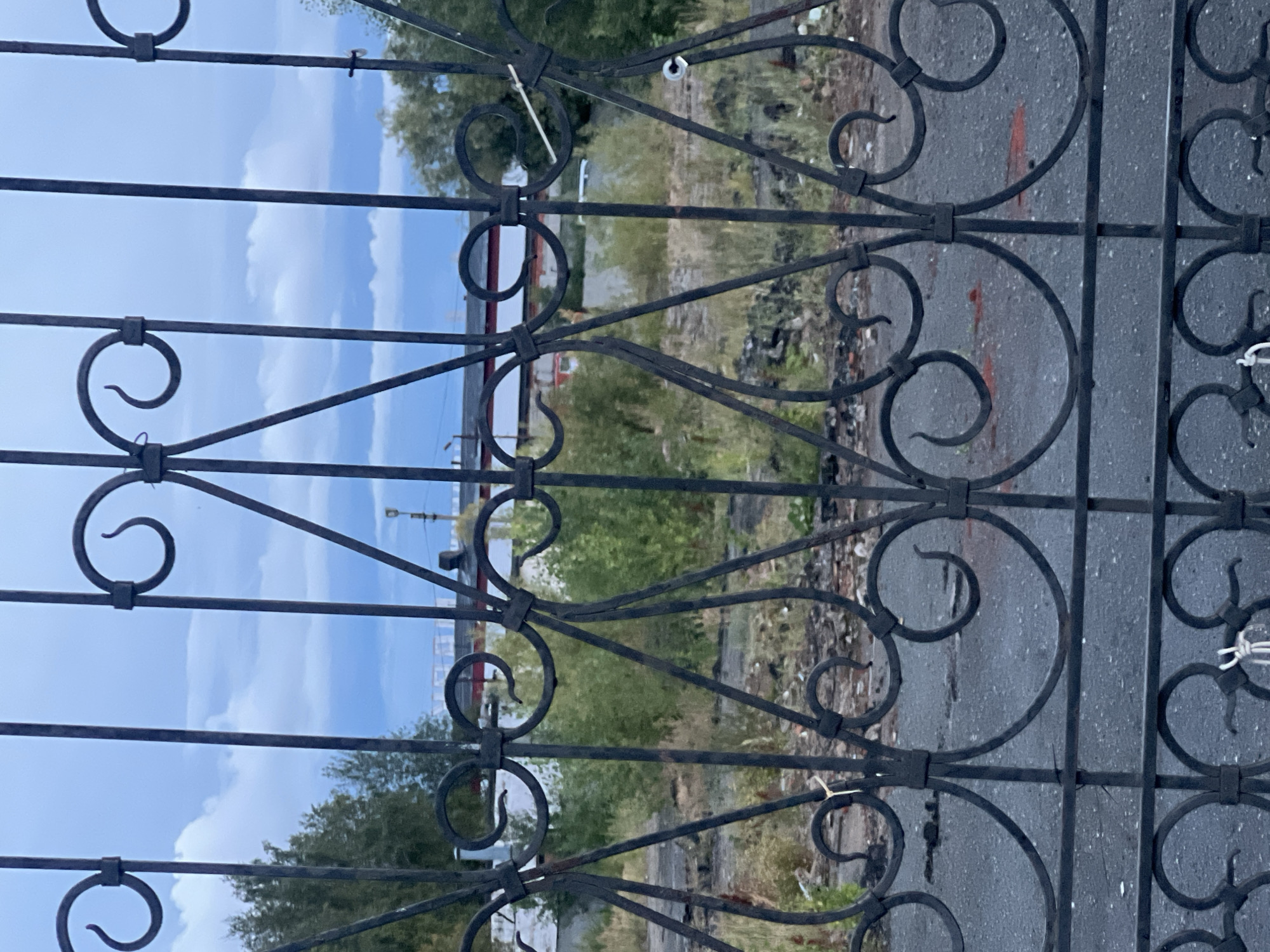 A close-up, eye-level shot, taken through an ornate, black metal fence, depicts a dimly lit, somewhat desolate industrial or abandoned lot. The fence itself, made of wrought iron, features a repeating pattern of vertical bars, curved decorative elements, and diagonal supports, with visible signs of wear and rust. Some white zip ties are attached to the fence.
Through the fence, the background reveals a gray, wet asphalt surface, possibly a road or parking lot, with scattered debris including dark rubble and some reddish-brown objects that may be bricks or tiles. Sparse, dry grass and green bushes are growing in patches across the lot. Further back, a low-rise building with a dark roof and white walls, accented with red, is visible. Beyond the building, a stand of green trees can be seen against a cloudy, overcast sky.
The overall impression is one of neglect and disuse. There are no people or animals visible in the frame, and no active events are taking place. The scene suggests an outdoor location in an urban or semi-urban environment. Given the provided context of Surgut, Russia, this could be an area near industrial facilities, a former factory site, or a storage yard.