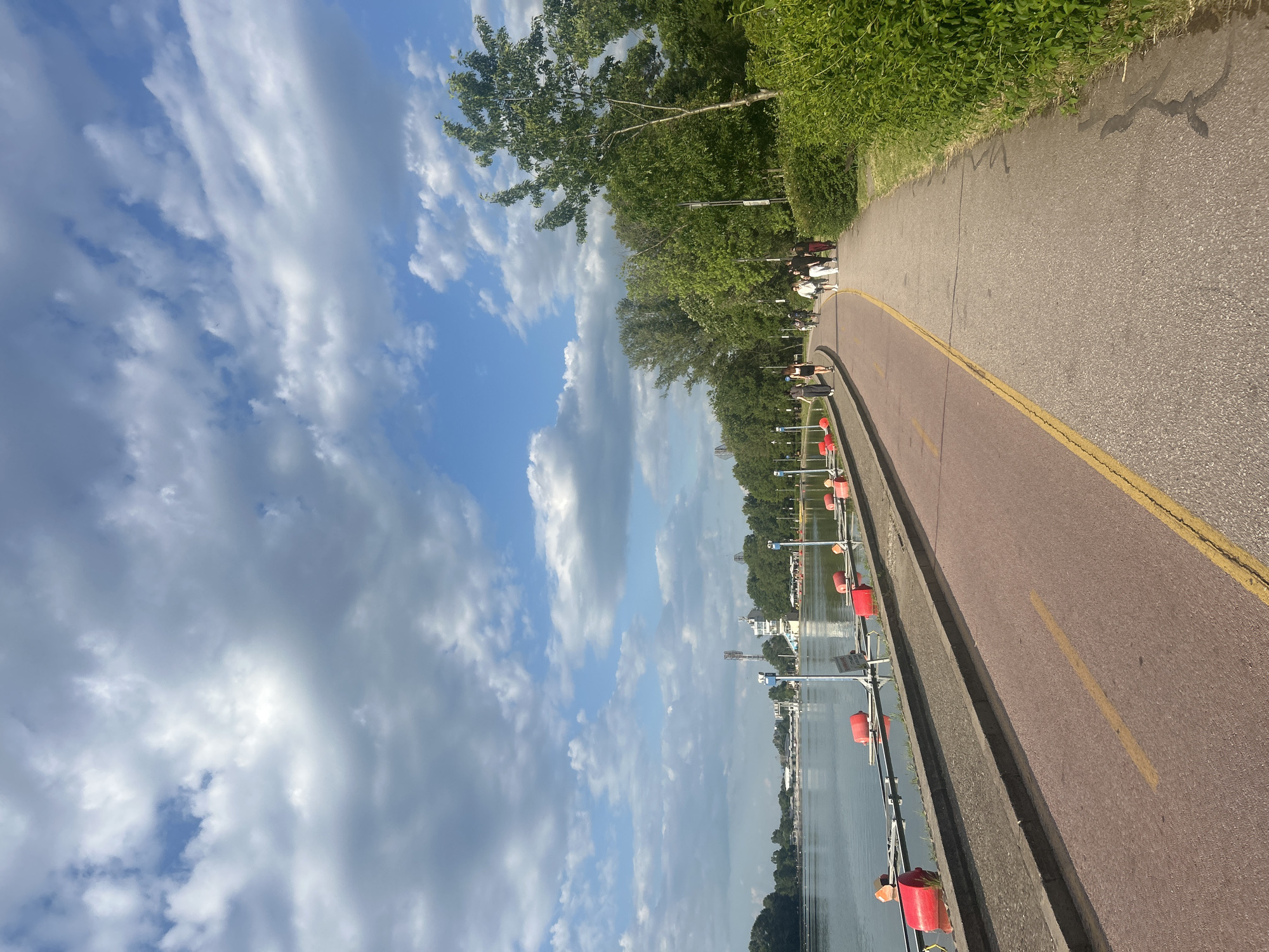 The image shows a lakeside scene in Plovdiv, Bulgaria. A paved pathway runs along the edge of a lake, bordered by a concrete barrier and a line of red buoys. Trees line the pathway on one side. In the distance, buildings and structures are visible on the opposite side of the lake. The sky is partly cloudy. People walk along the pathway.