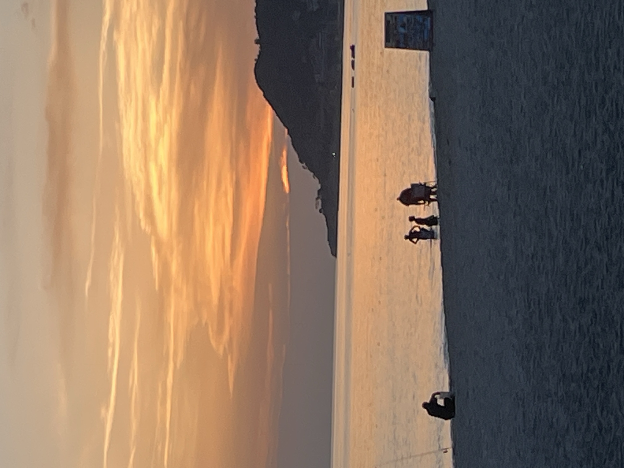 The image is a sideways view of a beach at sunset, likely in Alanya, Turkey given the visible mountainous silhouette. A family consisting of two people and a baby sits in a stroller at the center of the beach. A person is casting a fishing pole at the edge of the beach. The sky is a blend of orange and yellow hues, indicating a sunset or sunrise. The water is calm and reflects the colors of the sky. A sign is visible in the upper right corner. The beach itself appears sandy.