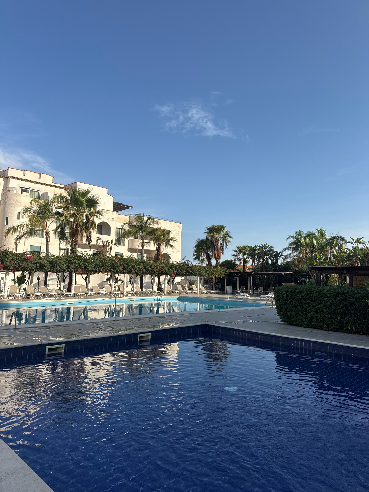 The image depicts a resort setting with a swimming pool in the foreground. The pool's water is a deep blue and reflects the surrounding environment. In the background, a multi-story building with white walls and arched windows is visible, adorned with palm trees and flowering vines. Adjacent to the pool, lounge chairs are arranged under a pergola. The scene suggests a sunny day with a clear blue sky and a few scattered clouds. The location is specified as Isola delle Femmine, Italy.