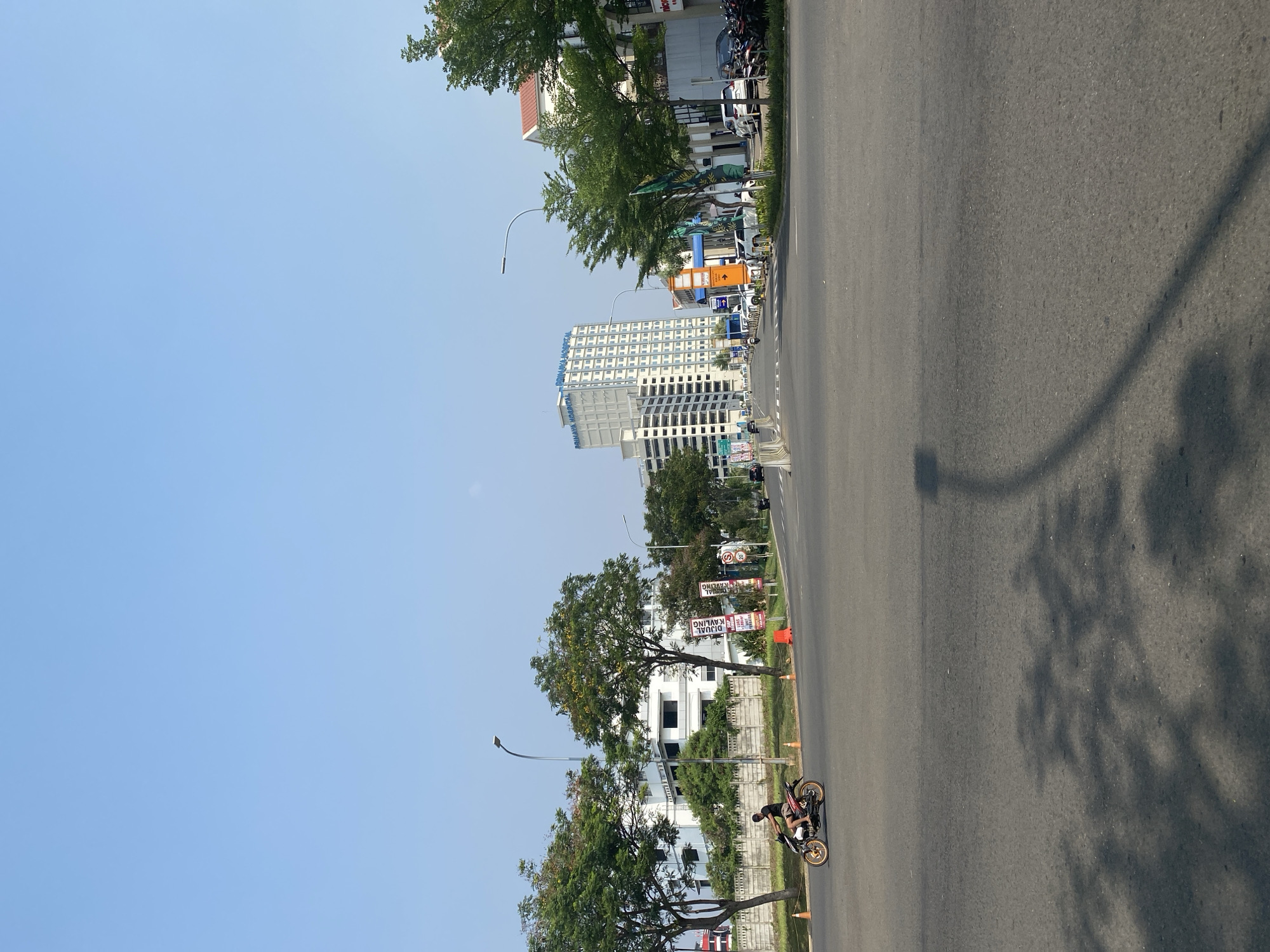 The image depicts a street scene in North Jakarta, Indonesia, under a clear blue sky. In the foreground, a paved road is visible with tire tracks and the shadows of trees cast across it. A person is riding a motorcycle with gold-colored rims on the left side of the road, heading away from the camera. Traffic cones are placed along the side of the road.
In the middle ground, several trees with green foliage line the street. Behind the trees, there are buildings. A tall, white, multi-story building with many windows stands prominently. Further down the street, other buildings and parked vehicles are visible. Signs with text, some in Indonesian, are also present. The environment appears to be an urban setting, likely a commercial or residential area.