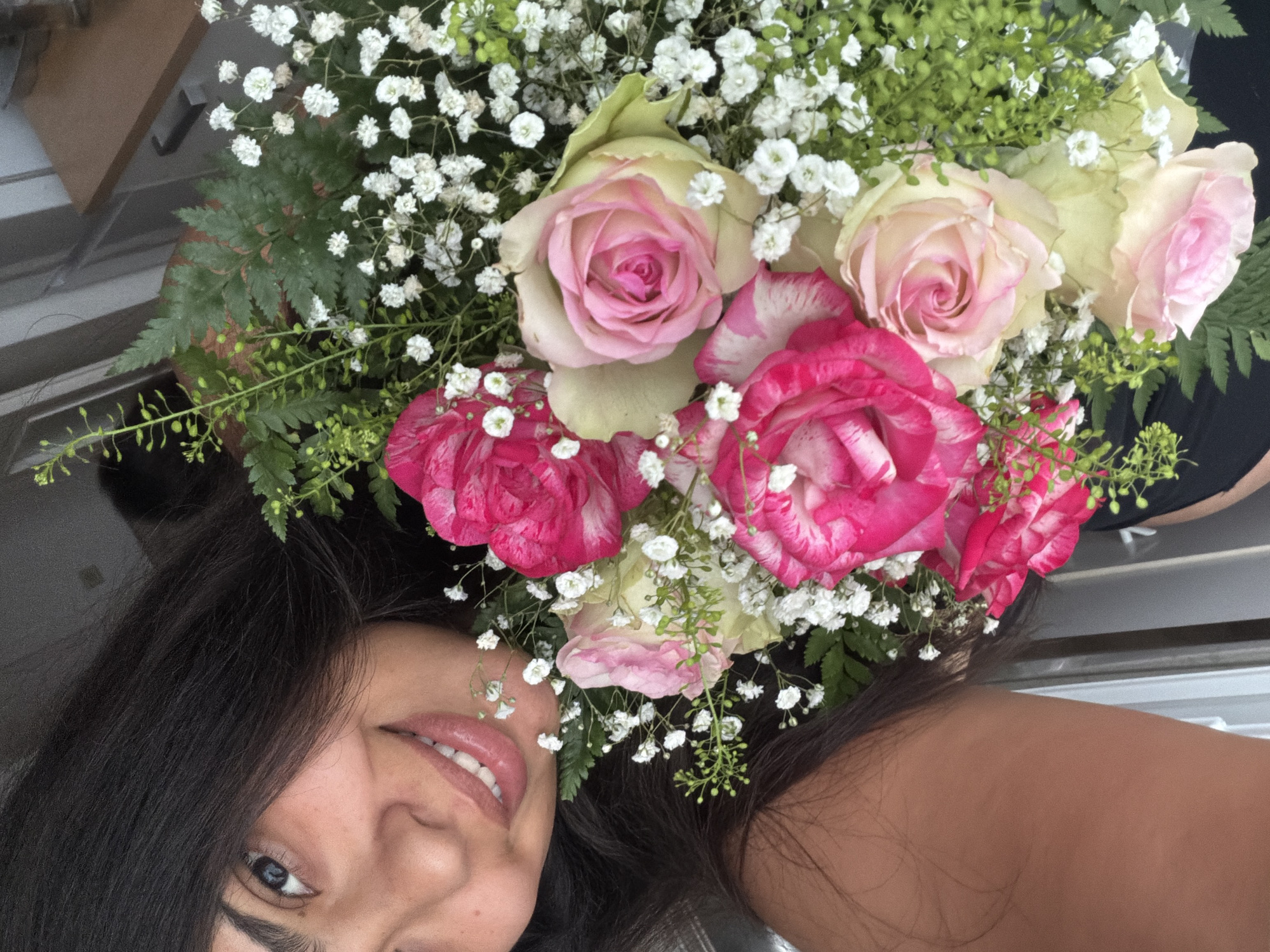 The image is a close-up, selfie-style photograph of a young woman holding a bouquet of roses and filler flowers. She is smiling, with her left eye visible and her mouth open in a grin. Her dark hair is falling forward, partially obscuring her face. The bouquet contains pink and white roses, along with small white flowers and green foliage. The background is indistinct, with hints of an indoor setting possibly including furniture and a window. The image is taken from a slightly elevated angle, looking down at the woman. There is no discernible information about the location within the city of Duisburg, Germany.