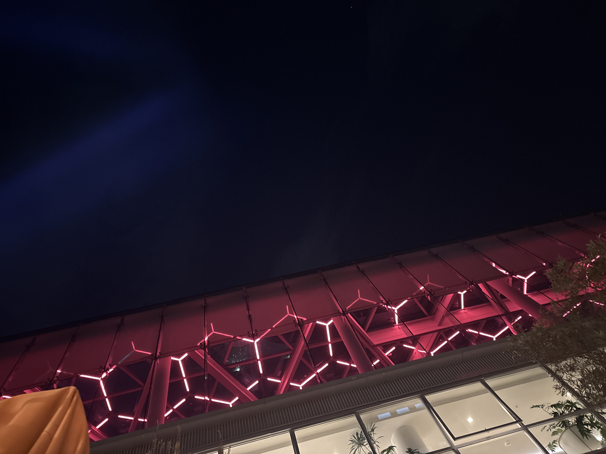 The image is a nighttime shot of a building in Mexico City, Mexico. The building has a unique architectural design, featuring a glass facade and a geometrically patterned roof illuminated with red lights. Below the roof, there are large windows offering a glimpse into the interior, where plants are visible. The sky is dark, with a hint of blue in the upper left corner. Some foliage is seen on the right side of the frame.