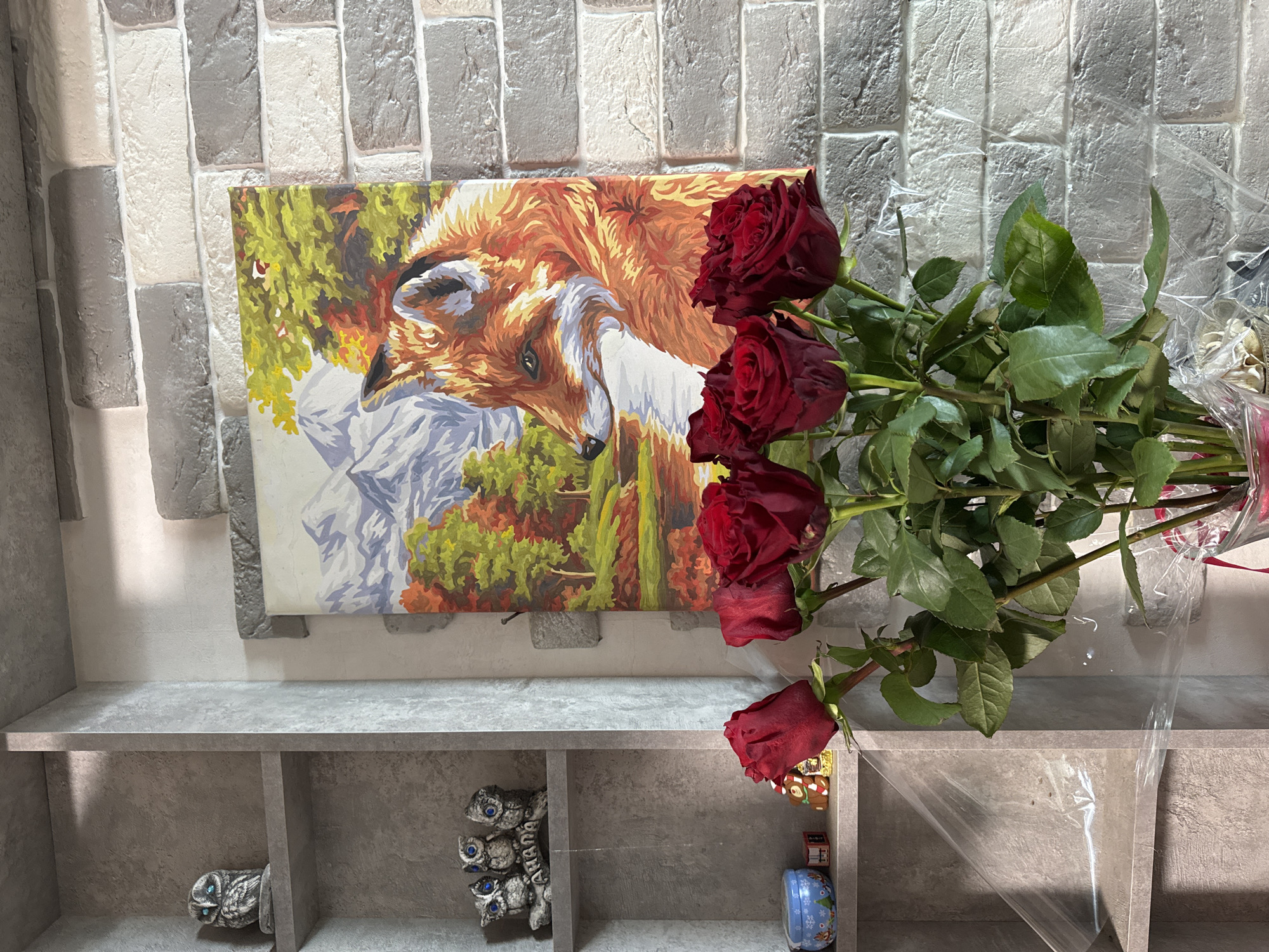 In Chaikovsky, Russia, a still life photograph captures a bouquet of deep red roses in the foreground, their petals partially unfurled and leaves a vibrant green. Behind the roses, a painting is displayed on a textured, light-gray brick wall. The artwork depicts a fox with its head turned to the side, set against a backdrop of autumnal trees and snow-capped mountains. To the left of the painting, a gray shelving unit contains decorative owl figurines on its upper shelves. The overall environment appears to be indoors, possibly a home or a studio, given the arrangement of the objects.