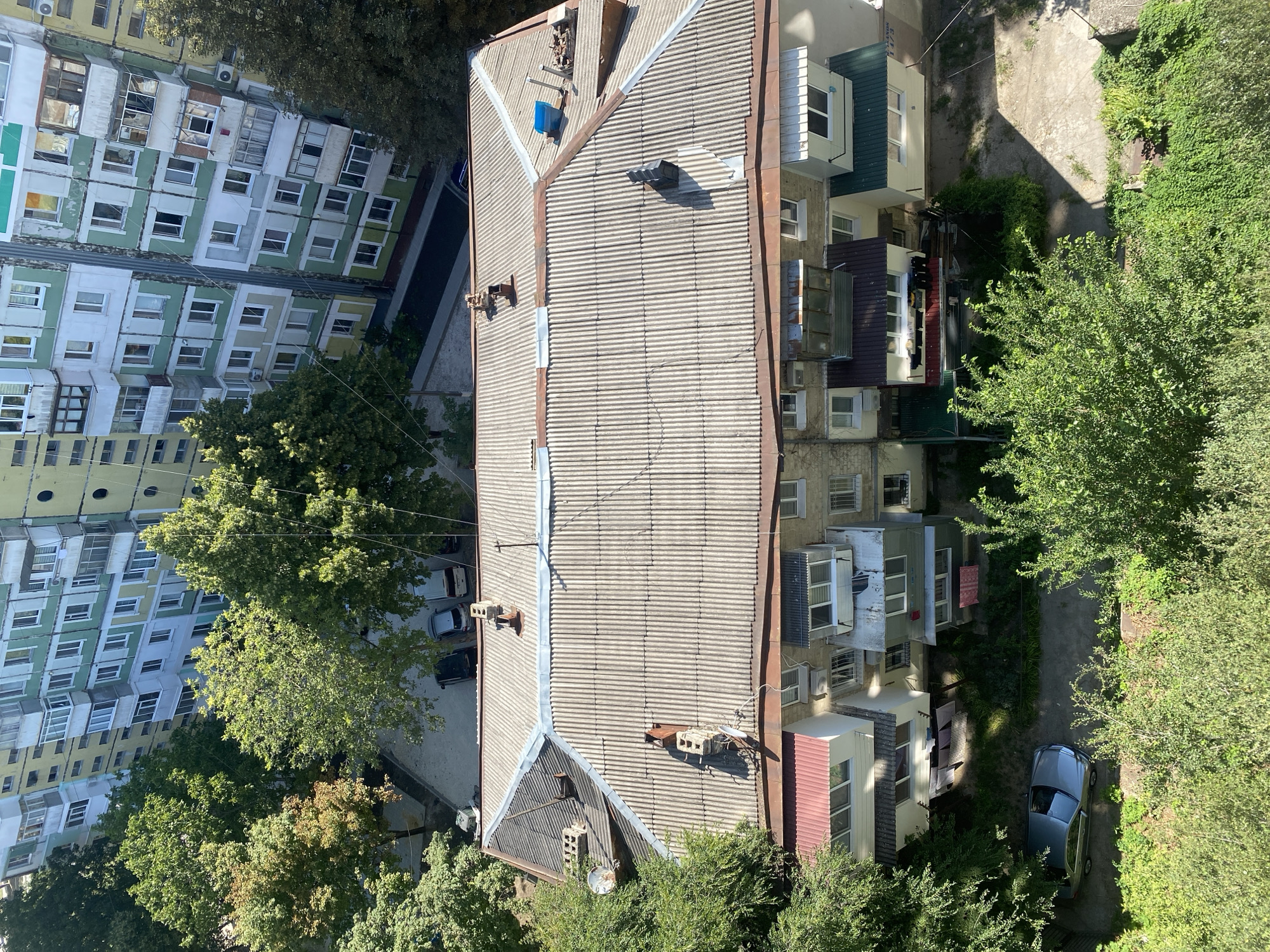 This is an aerial view of a residential building in Chisinau, Moldova. The building is multi-story with a light-colored, corrugated roof. Several chimneys and ventilation pipes are visible on the roof, some of which appear to be constructed from brick. A satellite dish is mounted on the roof of a smaller section of the building.
The main building has a light grey, corrugated roof, with some sections appearing weathered or discolored. The facade of the building is a mix of cream and light green, with numerous windows and balconies. On the ground level, there is a small paved area with a silver sedan parked. Surrounding the building are lush green trees, some with developing autumn colors, and dense bushes. In the background, another larger, multi-story apartment building is visible, featuring a colorful facade with alternating bands of yellow and green, and many windows and balconies. The environment appears to be a typical urban residential area. There are no people visible in the image.