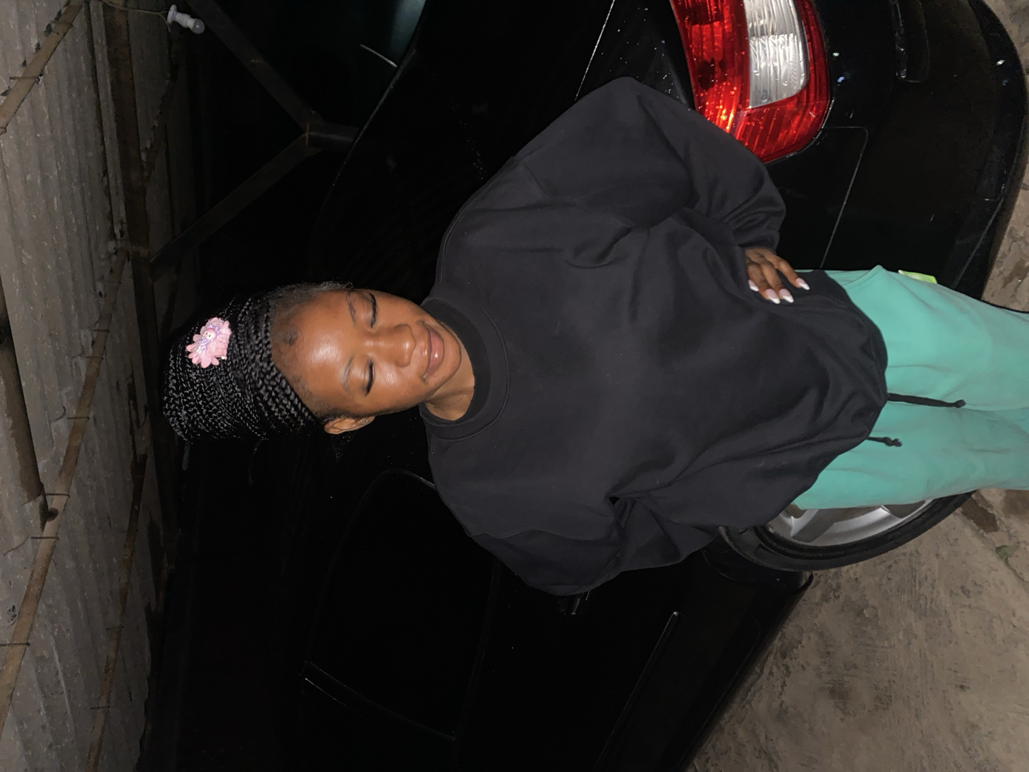 A young Black woman with braided hair adorned with a pink flower clip stands with her arms behind her back in front of a black car. She is wearing a black sweatshirt and teal sweatpants. The scene appears to be in an outdoor, covered area, possibly a garage or carport, with a concrete ceiling visible above. The ground is also concrete. The lighting suggests it is nighttime or indoors with artificial light. The image is taken in Jos, Nigeria.