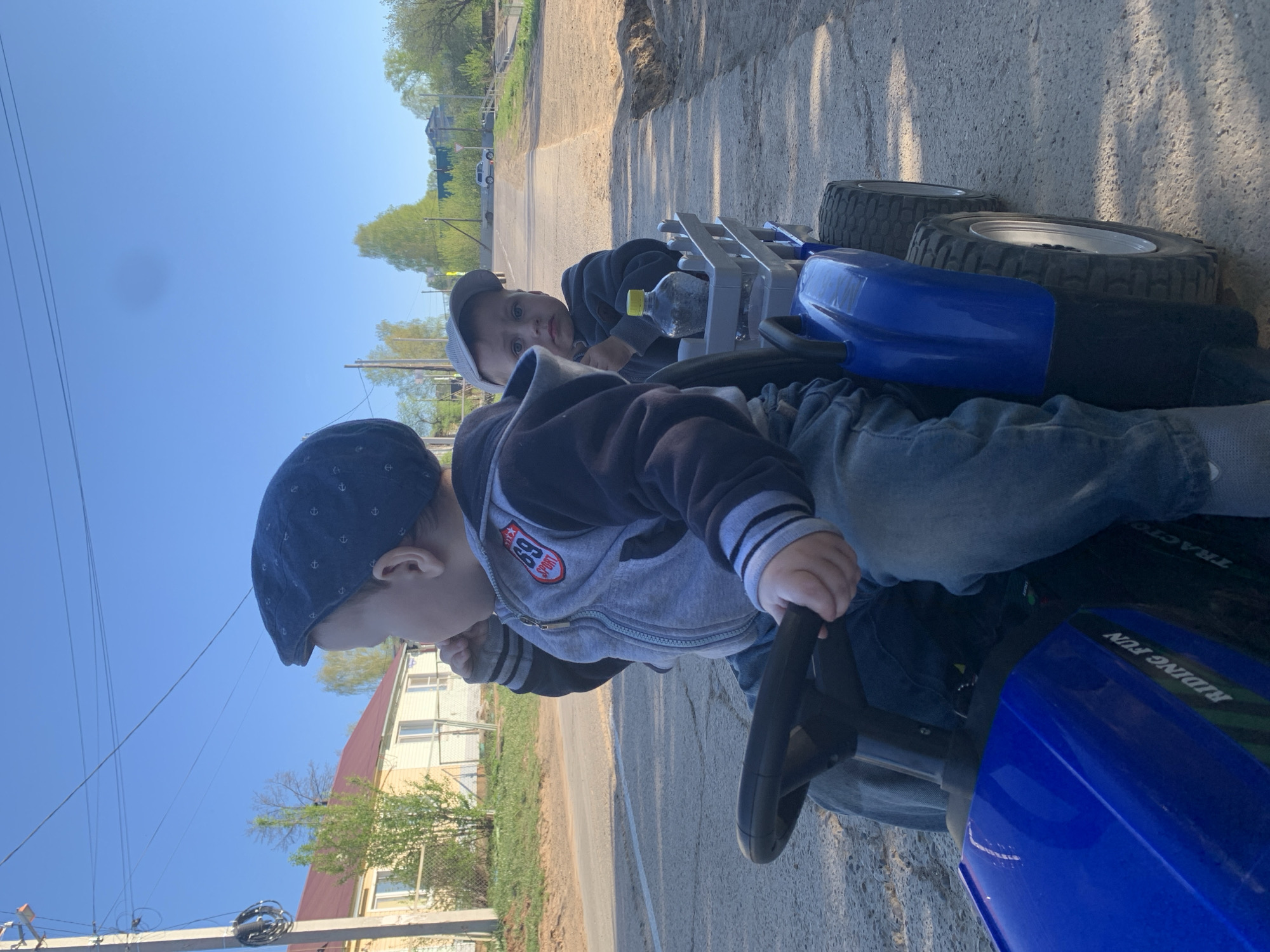 The image shows two young boys riding a blue toy tractor on a paved road. The older boy is driving, wearing a dark cap, gray and black jacket, and jeans. The younger boy sits behind him, also wearing a cap and dark clothing. A clear water bottle rests in the tractor's storage rack. The road runs through what appears to be a residential area in Фоки, Russia. Houses and trees line the street, with utility poles and wires visible overhead against a blue sky.