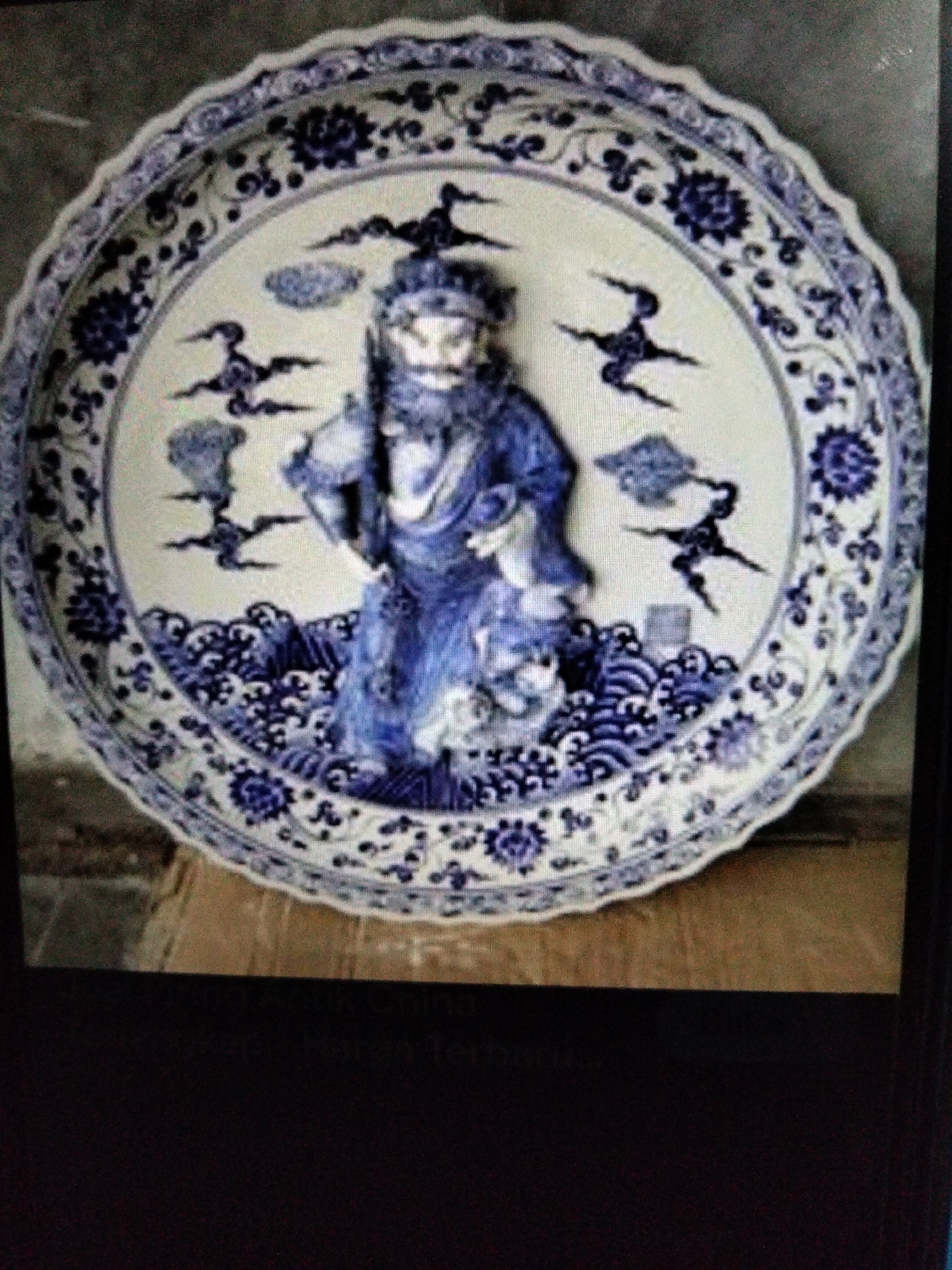 The image shows a large, decorative ceramic plate with a scalloped edge, adorned with blue and white patterns. The central motif features a figure in traditional blue attire, possibly a deity or historical character, with a beard and elaborate headdress. The figure is depicted standing amidst swirling waves and stylized clouds. The rim of the plate is decorated with floral and geometric designs in blue. The plate is placed on a wooden surface, possibly a table or a shelf, with a textured background. No specific details about Kualaraja, Indonesia are visible or inferable from the image.