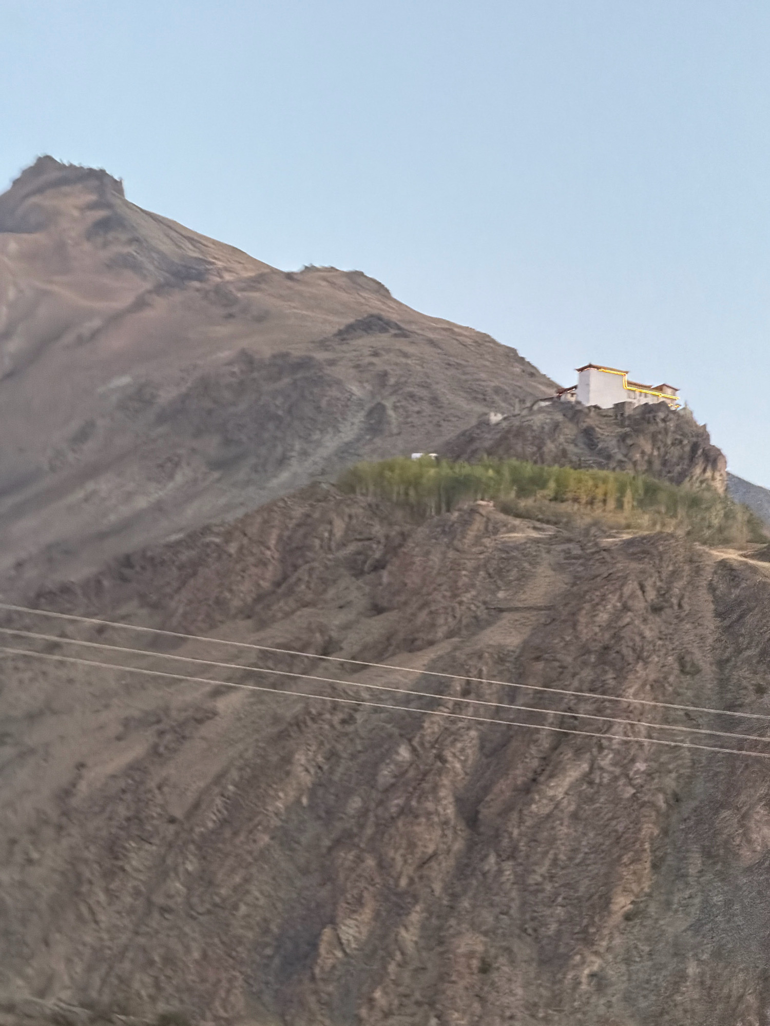 The image shows a monastery perched on a rocky mountain slope in Stongde, India. The monastery complex, with its white walls and yellow accents, stands out against the brown and gray tones of the mountainous terrain. A cluster of trees, possibly planted, are visible on a lower terrace below the monastery. The sky above is a clear, pale blue, indicating fair weather. Three thin, parallel wires, likely power lines, traverse the lower part of the image, indicating human infrastructure in the otherwise natural landscape. The overall scene suggests a remote, high-altitude location.
