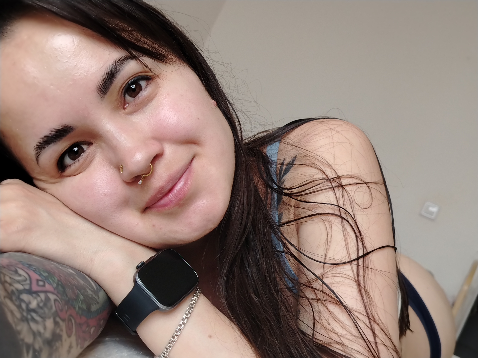 A woman with brown hair is resting her head on her left arm, with her right arm also visible. She has dark eyes and eyebrows, a nose ring, and is smiling slightly. She is wearing a smart watch on her left wrist, along with a silver bracelet. A tattoo is visible on her left arm. She is wearing a blue top. The background is a plain, light-colored wall. The location is Unknown, Unknown.