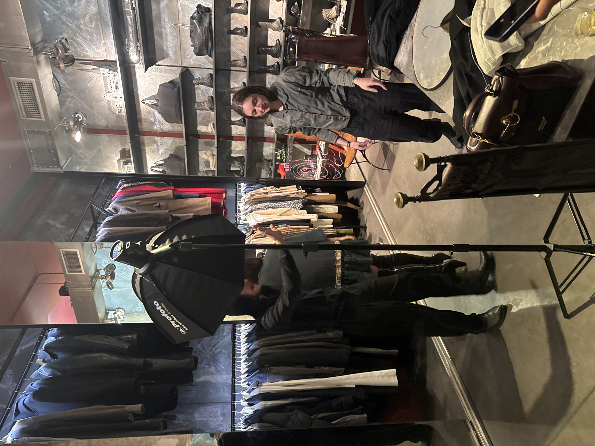 The scene appears to be an upscale retail store, likely in Paris, France, featuring high-end fashion items. Shelves laden with handbags and footwear line one side of the shop, while racks of clothing, including jackets, are on the other. Two individuals are present: One person is seated near a display of shoes and handbags, wearing a light-colored jacket and dark pants. Another is positioned near a clothing rack and is obscured by a softbox lighting device on a stand. A dark brown handbag is prominently displayed on a nearby surface. The environment suggests a chic, modern aesthetic typical of luxury retail spaces.