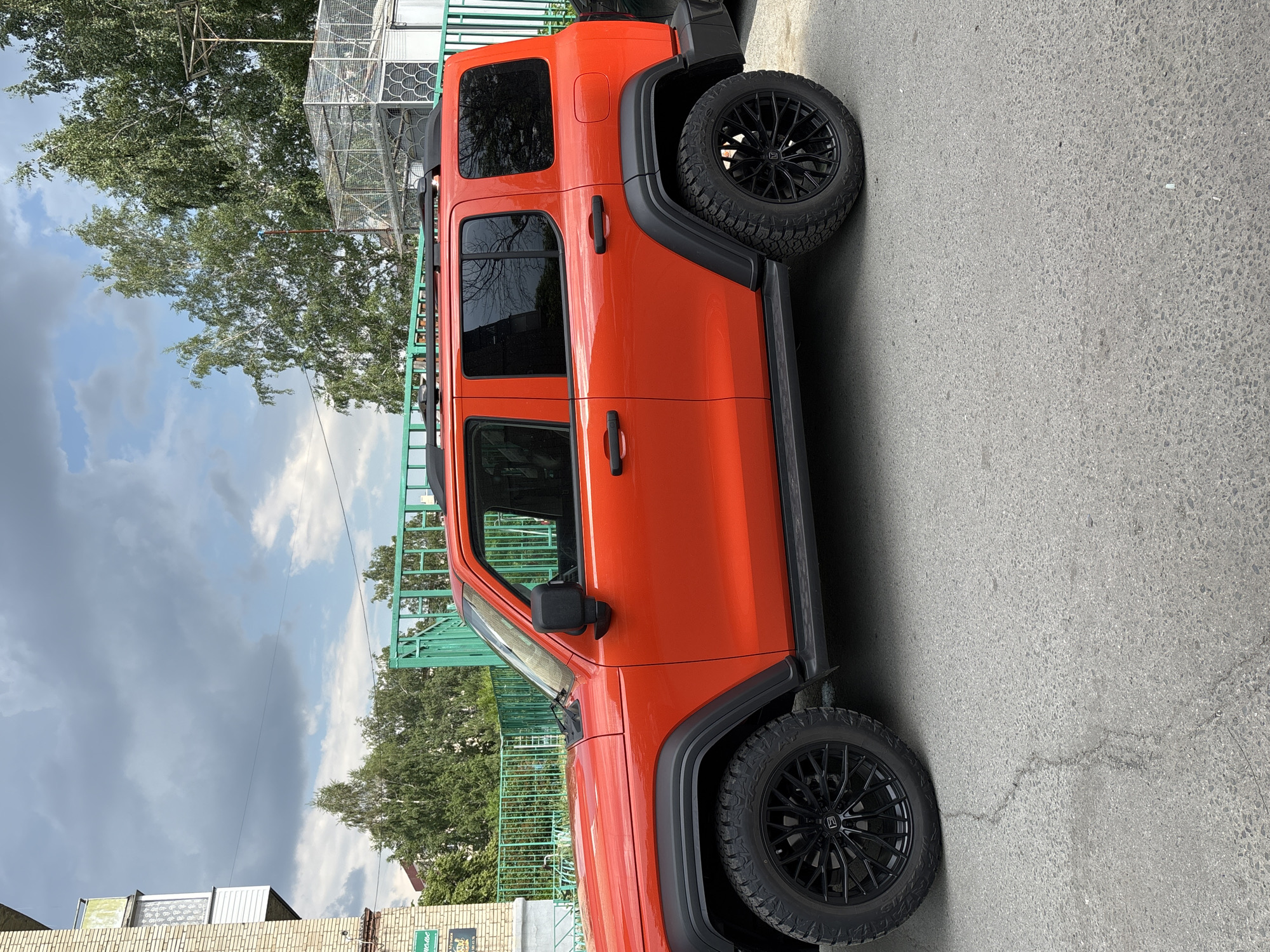 An orange SUV is parked on a paved street in front of a green metal fence. The SUV has black off-road tires and black roof rails. Behind the fence, there are trees and a building with a balcony. The sky is cloudy. The street appears to be in an urban or suburban area. The image was taken in Novosibirsk, Russia.