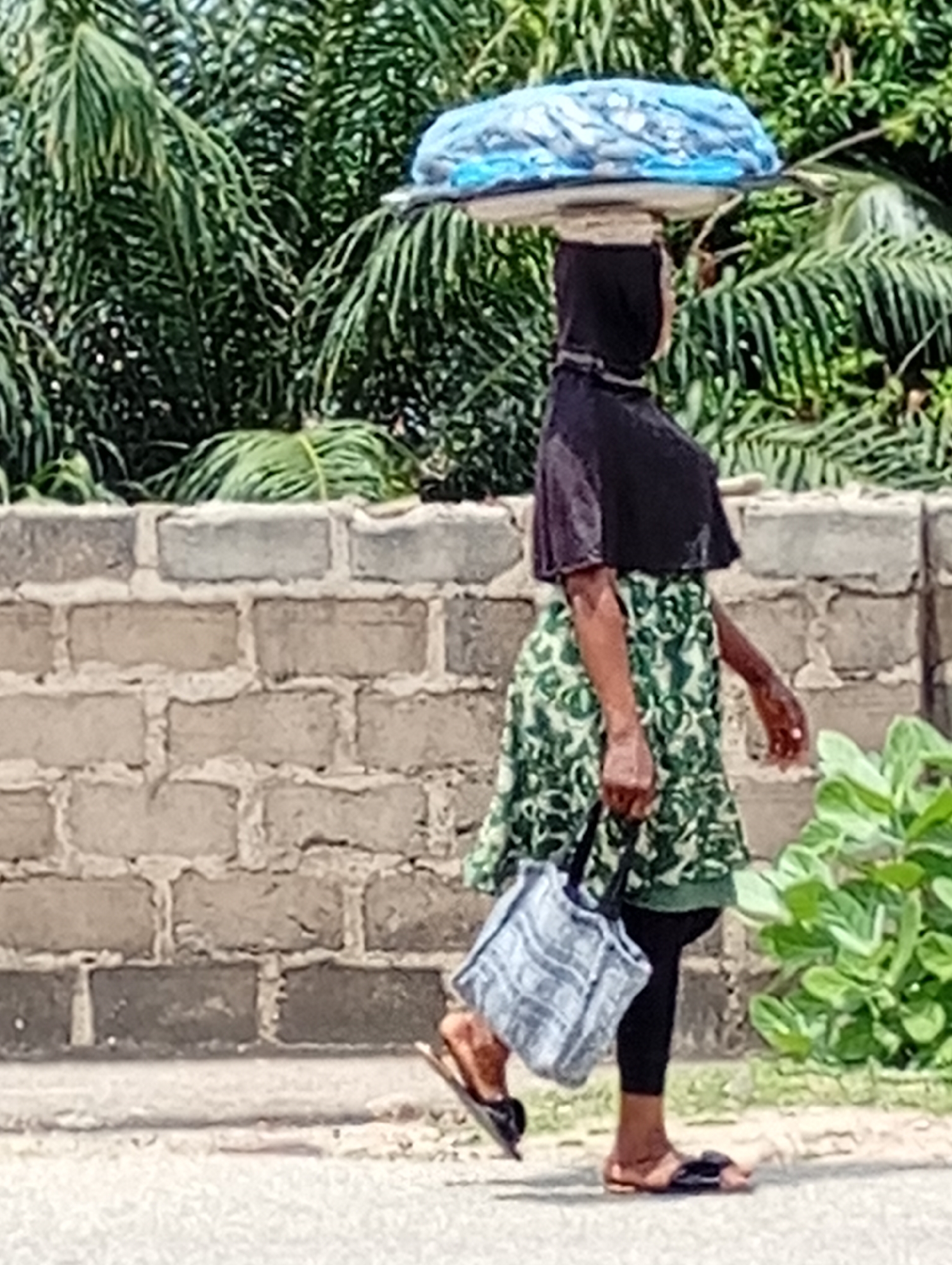 A woman is walking along a street in Lokoja, Nigeria. She has a large, blue-wrapped object balanced on her head, secured with a dark-colored head covering. She is wearing a short-sleeved, green and white patterned dress over black leggings, and brown sandals. She carries a light-colored, patterned shopping bag in her left hand. The background includes a low brick wall, lush green foliage, and a paved road. The scene is illuminated by bright, diffuse sunlight.