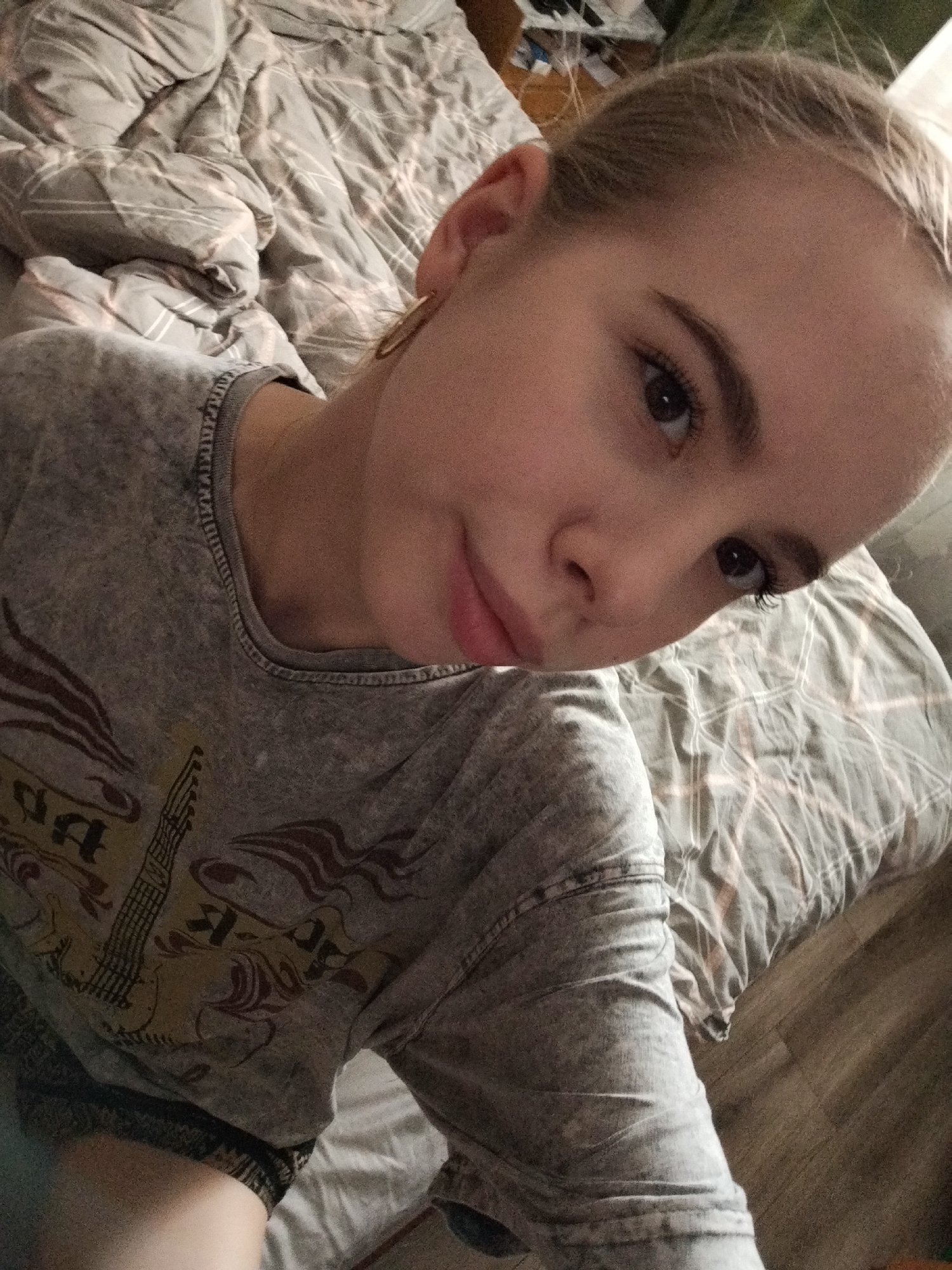 A young woman with blonde hair pulled back is seen in a selfie. She is lying on a bed with a grey, patterned comforter. The woman is wearing a grey t-shirt with a graphic print. She has on a hoop earring in her left ear. The lighting suggests an indoor setting, possibly a bedroom. The focus is on her face and upper body, with the bed and a glimpse of wooden flooring in the background.