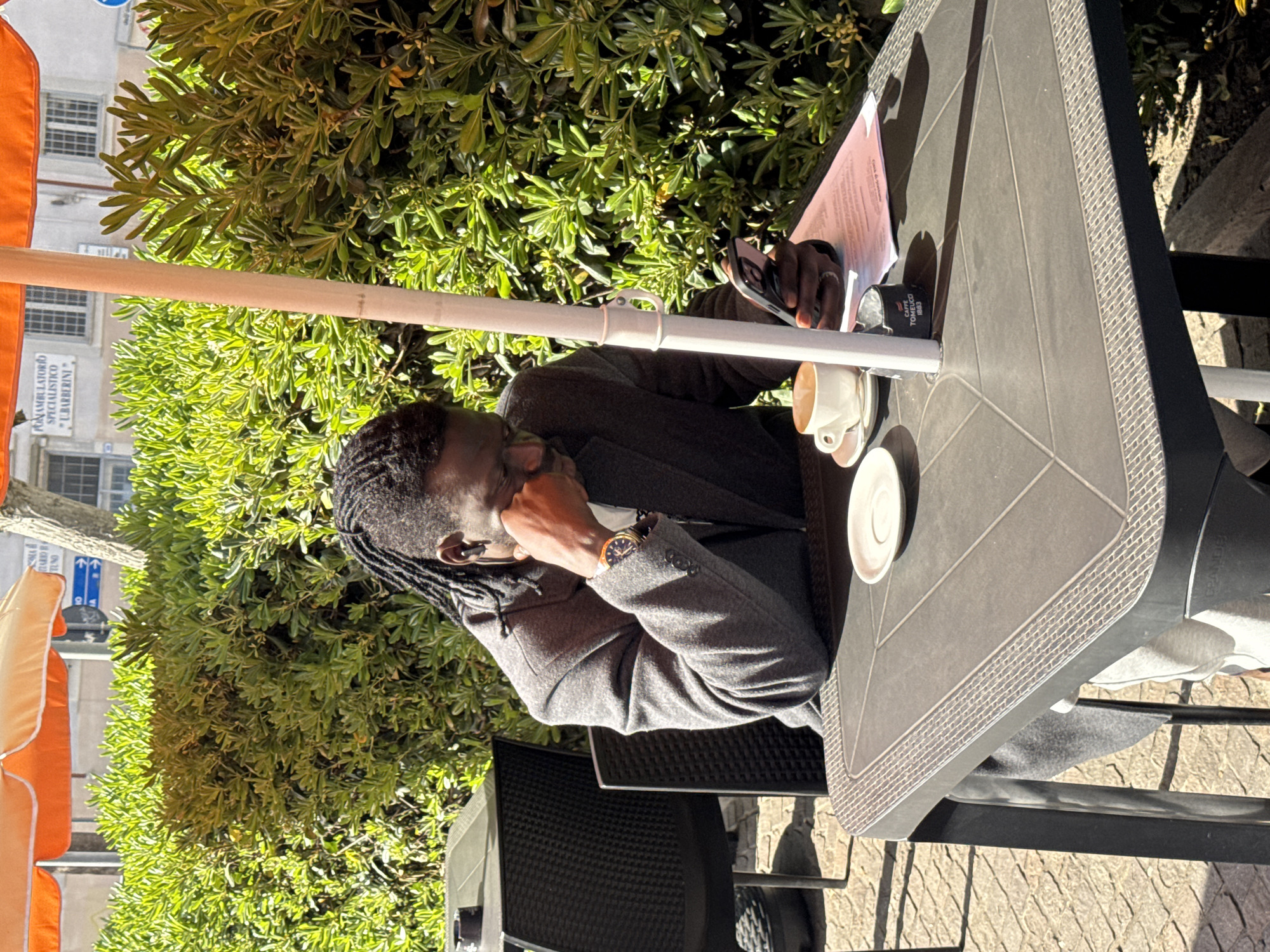 A man is sitting at an outdoor cafe table in Nettuno, Italy. He is wearing a grey coat and has dreadlocks. He is holding a phone in his hand and has a coffee cup on the table. There are documents on the table. The cafe is near a green bush and an orange umbrella. A building with signage is visible in the background, reading "Poliambulatorio Specialistico - L. Barberini." The street is paved.