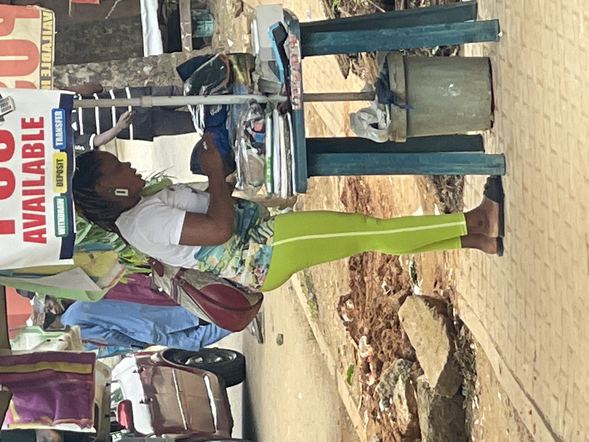 A woman stands at a stall in Abuja, Nigeria, examining items. She is wearing a white t-shirt with a colorful graphic, bright green leggings, and dark flip-flops. A maroon bag is slung over her shoulder. She is holding a small item, possibly a phone or a card reader, and looking down at it. The stall is covered by a banner advertising "AVAILABLE" services, with smaller text indicating "WITHDRAW," "DEPOSIT," and "TRANSFER." Behind her, another person in a blue traditional outfit is visible. The stall is laden with various goods, including folded clothes, books, and what appears to be a small white box and a blue device on a teal-colored table. A green cylindrical bin sits beneath the table. The ground is paved with interlocking bricks, and to the left, a red car is parked. The background shows a dirt embankment with rocks and debris.