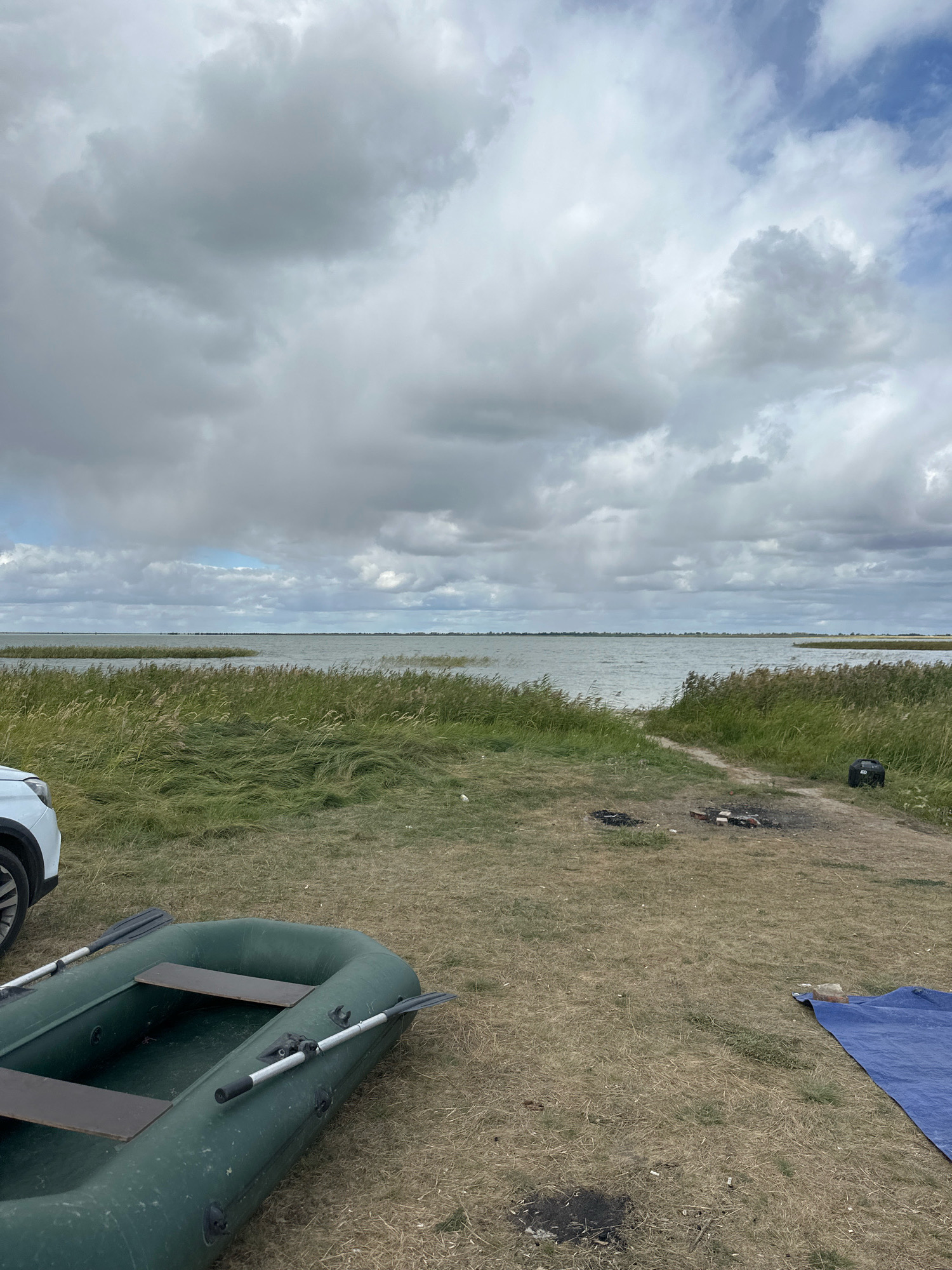 The image depicts a serene lakeside scene in Tsabelovka, Kazakhstan. In the foreground, a dark green inflatable boat with oars rests on dry grass. To the left, the front quarter of a white vehicle is visible. Further back, a path leads towards the water, bordered by tall, wind-swept reeds and grass. A small campfire pit with remnants of burnt wood is present on the path. In the middle ground, a vast, choppy body of water stretches to the horizon, under a dramatic sky filled with heavy, grey clouds with patches of blue sky peeking through. The water is lined with more reeds and vegetation on the far bank. To the right of the path, a black portable generator is placed on the ground, next to a folded blue tarp. The overall impression is one of a calm, natural setting, possibly a campsite near the lake.