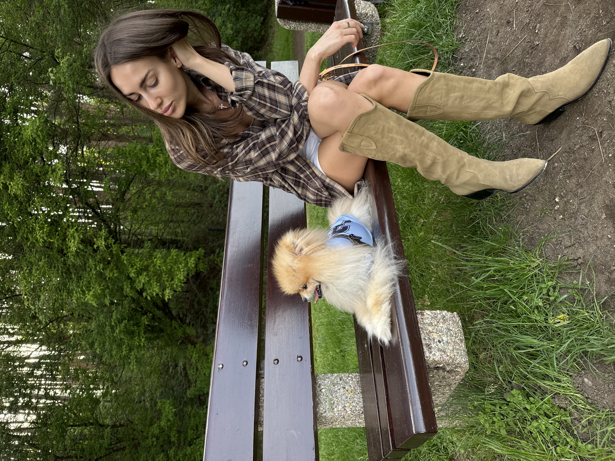 The image shows a woman and a dog on a park bench. The woman is seated on the bench, wearing a plaid shirt, light-colored shorts, and tan suede cowboy boots. She has long brown hair and is looking down with her hand near her head. A small, light brown Pomeranian dog wearing a blue vest is seated on the bench beside her. The bench is made of dark brown wood with silver rivets. The background includes trees and grass. The location appears to be a park or green space.