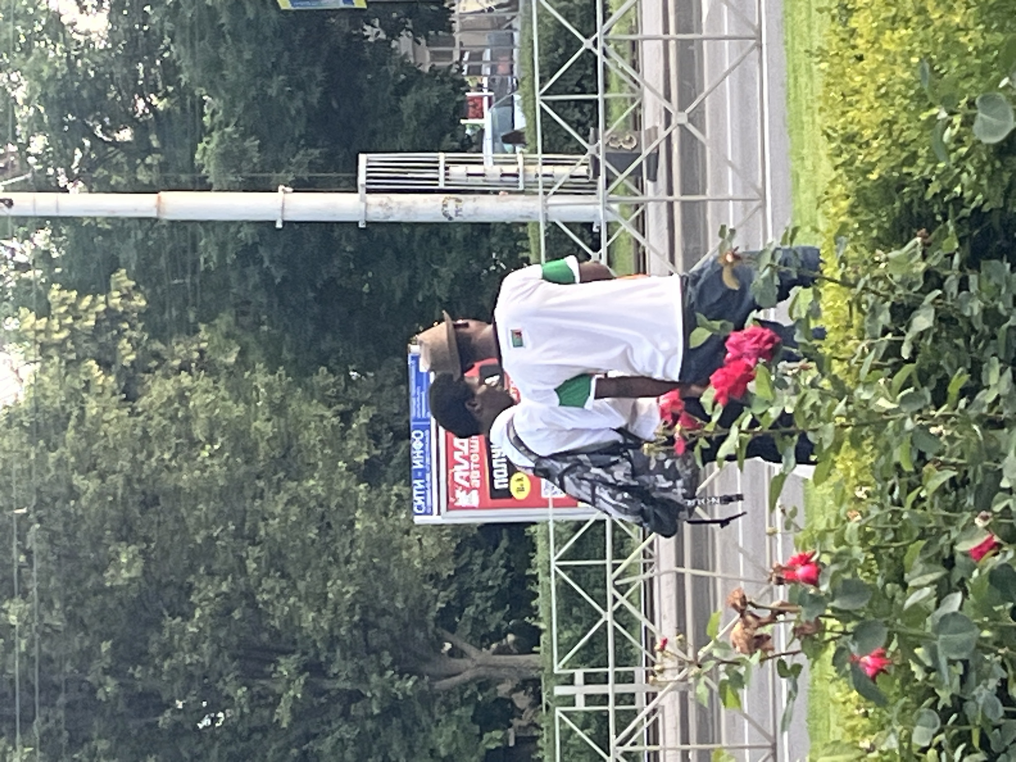 The image shows two young men standing by a street in Krasnodar, Russia. The man on the left is wearing a backpack and a white shirt. The man on the right is wearing a hat and a white shirt with green sleeves. They are standing in front of a fence, with trees and a billboard in the background. There are red roses in the foreground. The scene appears to be a street in the city.