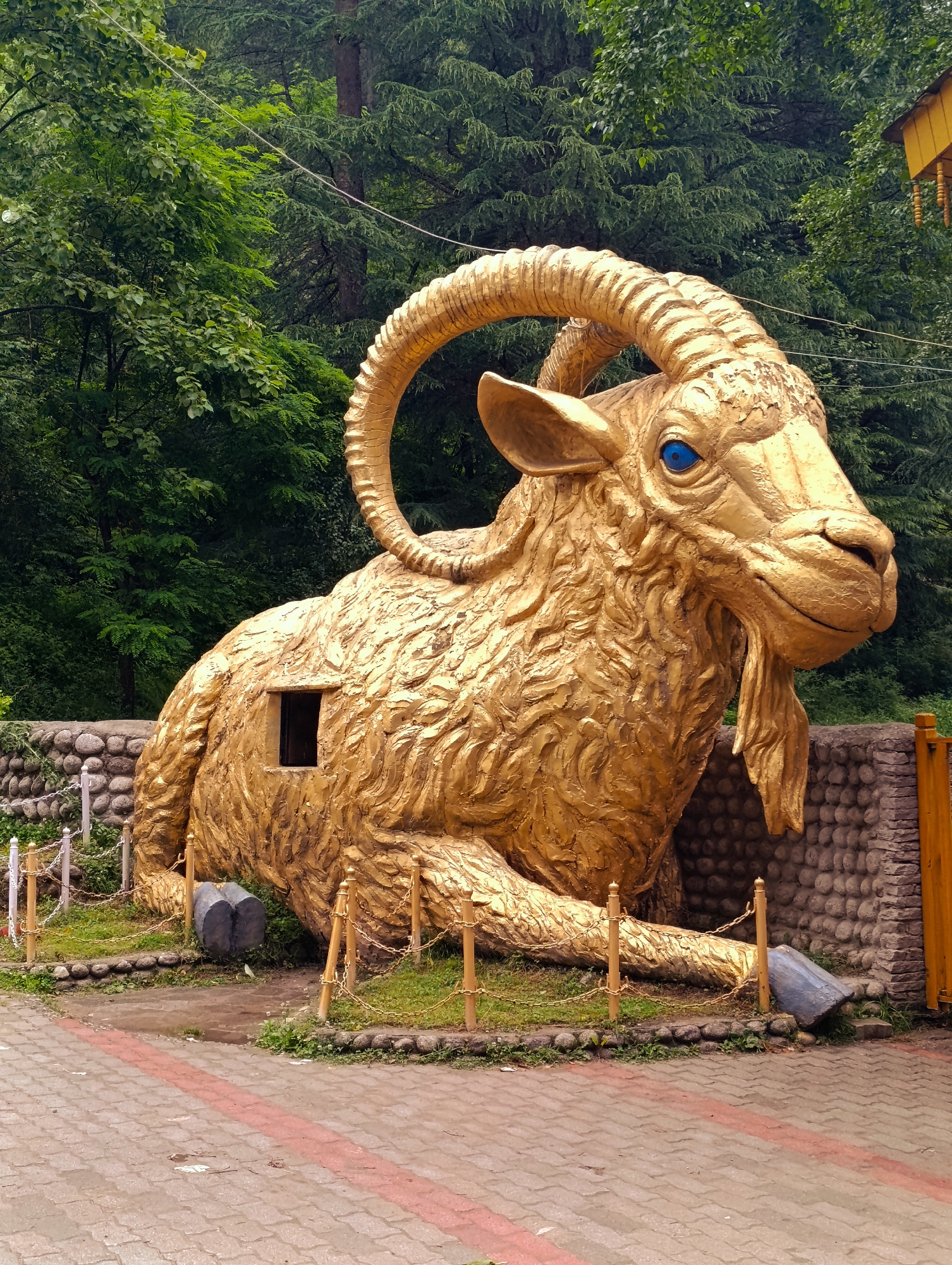 The image depicts a large, golden statue of a goat lying down, situated outdoors in Manali, India. The statue has bright blue eyes and large, curved horns. A small, dark rectangular opening is visible on the goat's body, suggesting it might serve as a small structure or exhibit. The statue is surrounded by a low wall made of stones, with a chain-link fence with wooden posts in front of it. Beyond the wall, a dense forest of green trees forms the background. The ground in the foreground is paved with interlocking bricks, with a red stripe running through it. There are no people visible in the image. The overall scene is that of an outdoor attraction or landmark in a natural, forested setting.