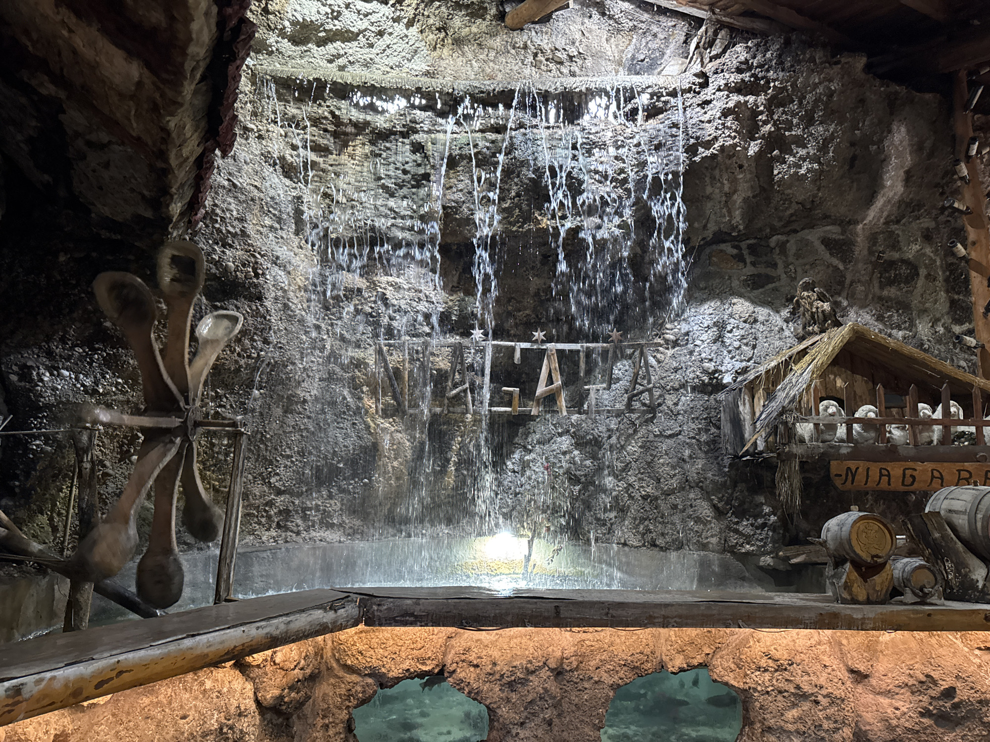 A photograph shows an indoor artificial waterfall exhibit in Podgorica, Montenegro. The waterfall cascades down a textured, rock-like backdrop into a pool of water. The word "NIAGARA" is displayed on a structure to the right of the waterfall, with miniature barrels nearby. A small wooden waterwheel is on the left. Fish can be seen in a pool beneath the structure that supports the waterfall. The scene is dimly lit.