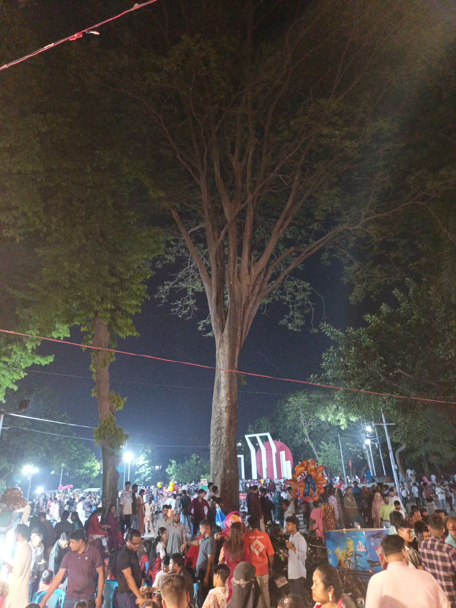 A nighttime scene shows a large crowd of people gathered at a park in Dhaka, Bangladesh. A tall, mature tree dominates the center of the frame, with other trees flanking it. Red electrical lines run across the top of the frame. In the background, the Shahid Minar monument is visible, indicating the location is likely near Dhaka University. People of various ages and attire fill the scene. Balloons and vendor stalls are also visible among the crowd. The lighting suggests it is nighttime, with spotlights illuminating the area.