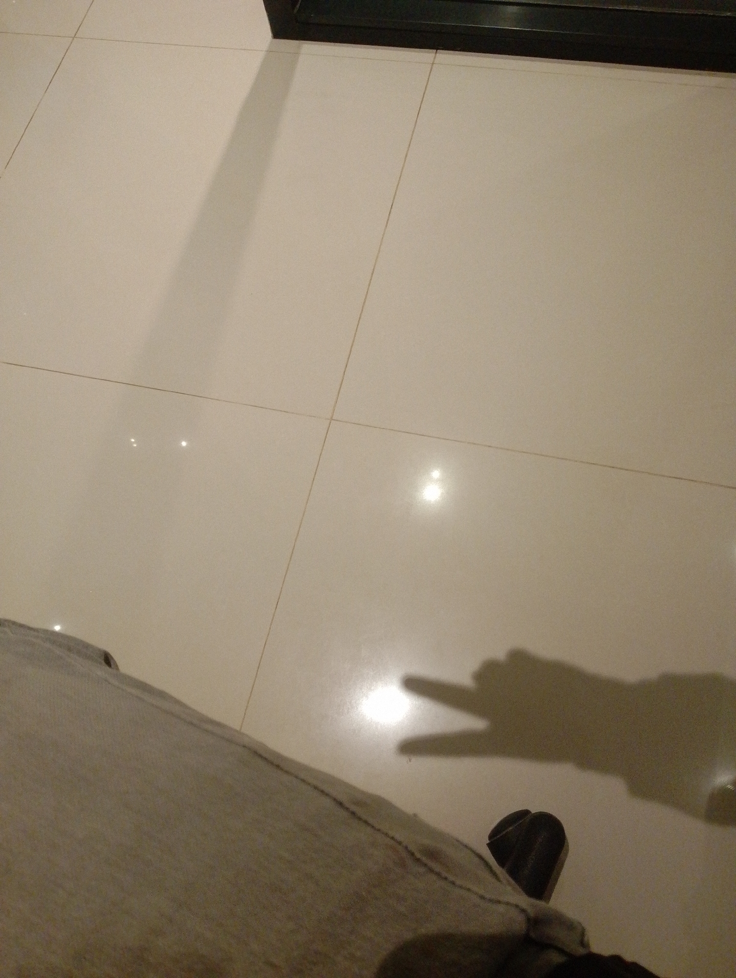 The image is a close-up, low-angle shot of a tiled floor. A person's legs, clad in light-colored pants, are visible in the foreground on the left, along with a black shoe. A shadow of a hand, making a peace sign, is cast on the floor to the right. The lighting suggests an indoor environment, possibly a commercial space, with reflections from light sources visible on the smooth, cream-colored tiles. There are no discernible landmarks or contextual elements to pinpoint the location specifically within Batulicin, Indonesia.