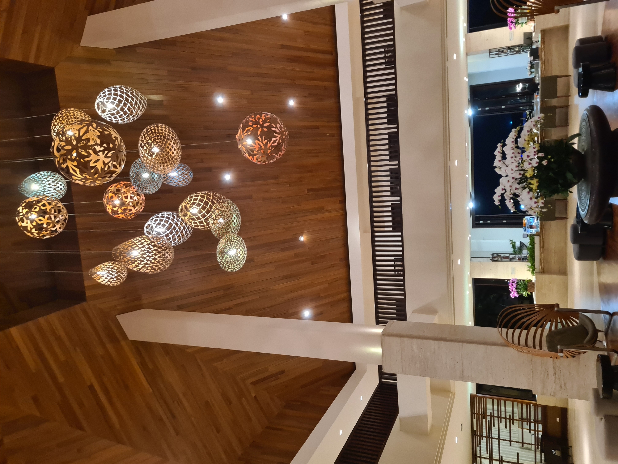 The image depicts the grand lobby of a hotel in Da Nang, Vietnam. A towering, multi-tiered chandelier made of ornate, circular metal globes with intricate cut-out patterns hangs from a high, wooden-paneled ceiling. The lobby features a large central pillar clad in textured stone, and a mezzanine level with dark wooden railings is visible in the background. In the foreground, a large, round, dark stone table holds a substantial arrangement of white orchids. The overall ambiance is luxurious and inviting, suggesting a high-end establishment.