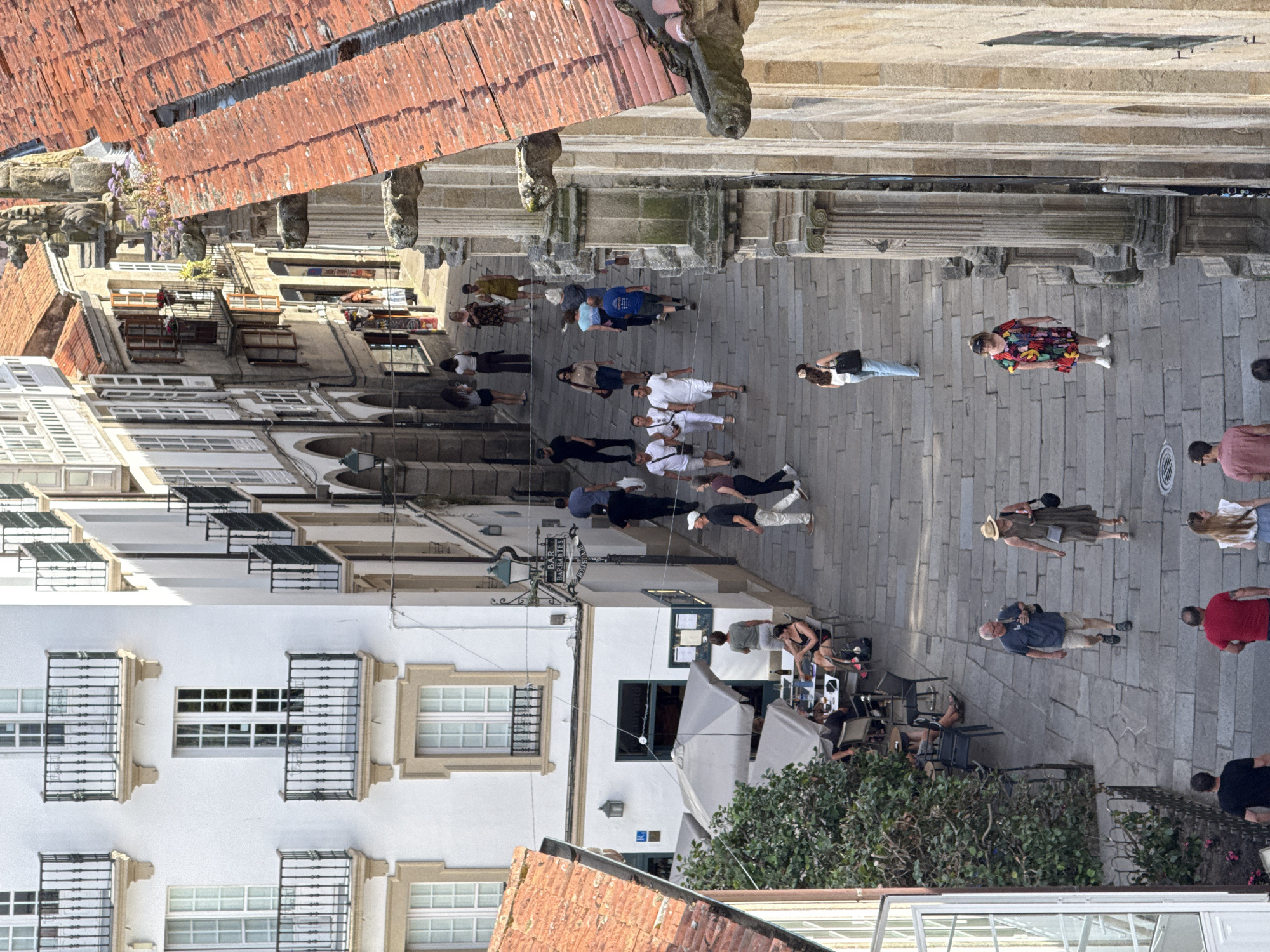 The image shows a street in Santiago De Compostela, Spain, featuring a steep stone stairway lined with historic buildings. On the left, a white building displays classical architecture with multiple windows and balconies. Adjacent to it, a narrower building features arcades on the ground floor and balconies on the upper levels. A restaurant with outdoor seating is visible under umbrellas.
On the right, a building with a red tiled roof and stone facade features detailed architectural elements, including columns and gargoyle-like figures. The stairway is populated by several people walking in both directions, some individually and some in small groups.
The overall scene depicts a lively urban environment with a blend of old-world architecture and contemporary pedestrian activity.