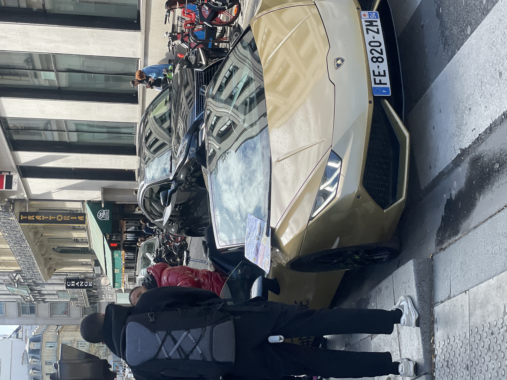 The image captures a vibrant street scene in Paris, France, characterized by a blend of historic and contemporary urban elements. In the immediate foreground, a gold-colored Lamborghini Huracán convertible is parked on the street, angled slightly towards the viewer. Its front license plate, "FE-820-ZM 94," is clearly visible, indicating a French registration from the Val-de-Marne department. A printed flyer or document rests on the vehicle's open soft-top roof, and small water droplets are discernible on its hood surface.
To the left of the Lamborghini, a person stands with their back to the camera, wearing a black hooded jacket, black trousers, and grey Nike sneakers. They are carrying a grey and black backpack with a distinctive criss-cross strapping pattern. Partially visible behind this individual and next to the Lamborghini is another person in a bright red puffer jacket.
Further down the street, directly behind the Lamborghini, a black Lexus SUV is parked. To the right of the Lexus, a rack of red public rental bicycles is present. Two additional individuals are observed in the midground, near a building facade on the right side of the street. One person has dark hair and is wearing a dark top, while the other, with reddish hair, wears a light blue top; both appear to be looking at mobile phones.
The surrounding environment consists of typical Parisian architecture: ornate, light-colored Haussmannian-style buildings with intricate balconies are visible on the left and in the background. A more modern building with large, dark-framed rectangular windows and a lighter facade stands on the right. Commercial signage is prominent, including a vertical neon sign displaying "CHANGE" and another for "TRATTORIA VESUVIO," an Italian restaurant with a green awning. The street pavement exhibits damp patches or puddles near the curb, and several cigarette butts are visible on the curb adjacent to the Lamborghini's front wheel. The scene is illuminated by bright daylight, suggesting clear weather.