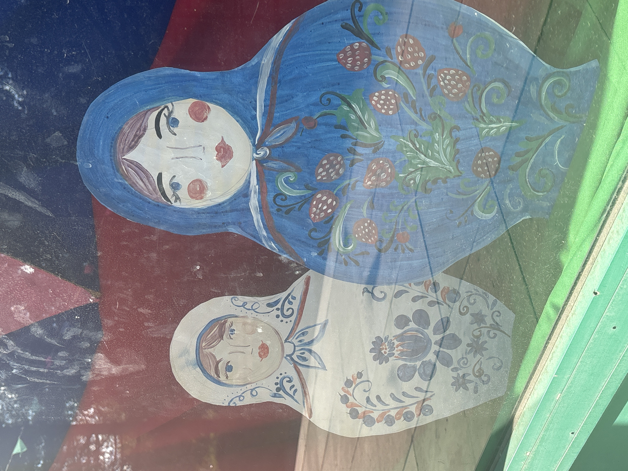 In the city of Ob, Russia, a close-up, eye-level shot captures a painting of two Matryoshka dolls on a window. The dolls are depicted in a traditional style, with the larger doll in the foreground painted in vibrant blue and adorned with a floral and strawberry pattern. The smaller doll, to the left and slightly behind, is painted in white with intricate blue and floral designs. The dolls are positioned against a backdrop that appears to be a painted mural featuring geometric shapes in red, blue, and black. The window glass shows reflections and some surface imperfections, suggesting it is part of a building's exterior. A bright green ledge runs along the bottom of the frame.